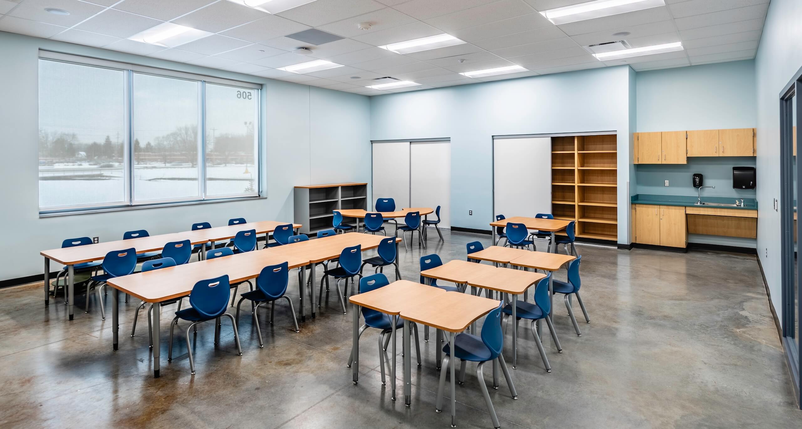 A modern classroom with blue chairs and wooden desks arranged in groups, bookshelves, a sink area, and large windows letting in natural light. The room has light blue walls and a polished concrete floor.