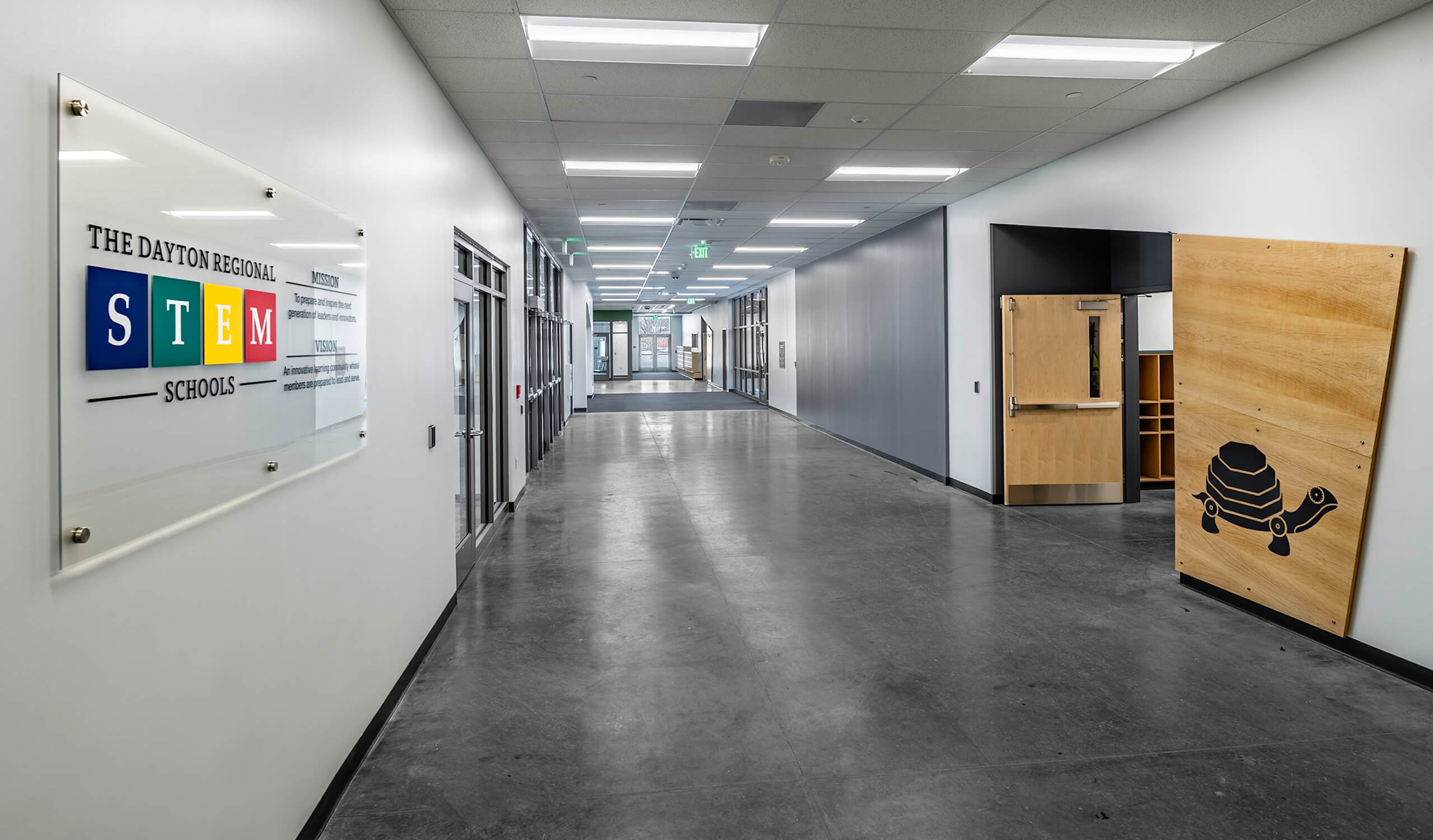 A clean, modern school hallway with gray floors, white walls, classroom doors, and a sign reading The Dayton Regional STEM Schools. A wooden panel with a robot graphic leans on one wall.