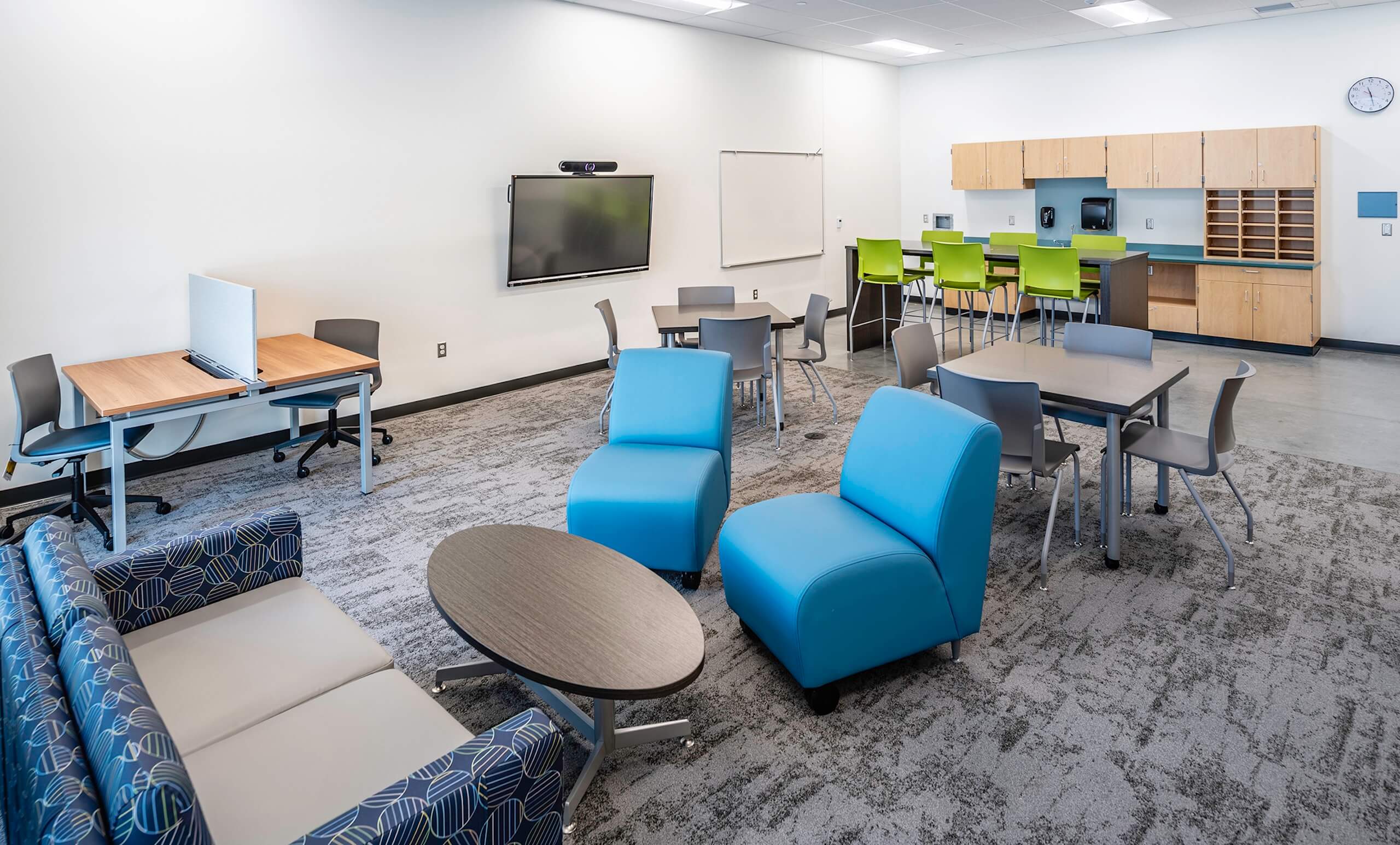 Modern classroom with blue and gray chairs, round tables, a sofa, a wall-mounted screen, a whiteboard, and a kitchenette area with green stools. The room has light walls and carpeted flooring.