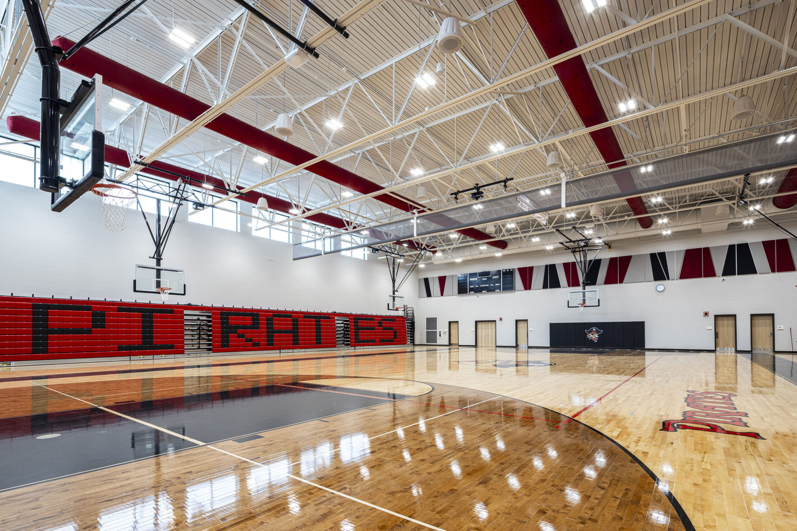 A high school gymnasium with polished wood floors, basketball hoops, red and black bleachers reading PIRATES, and geometric wall designs in red, black, and white.