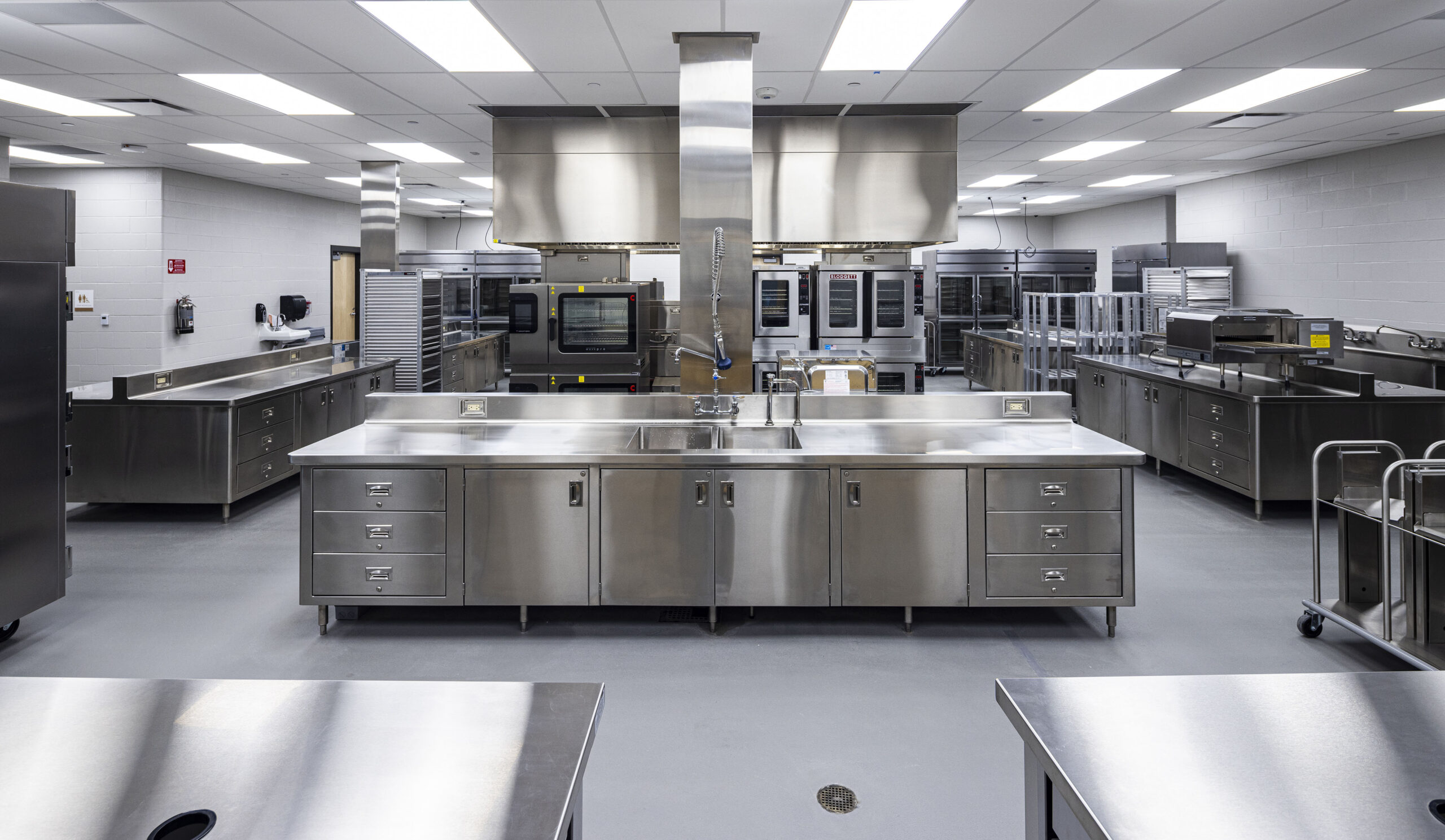 Modern commercial kitchen with stainless steel counters, appliances, ovens, and sinks. The space is clean, brightly lit, and organized, designed for large-scale food preparation or professional culinary use.