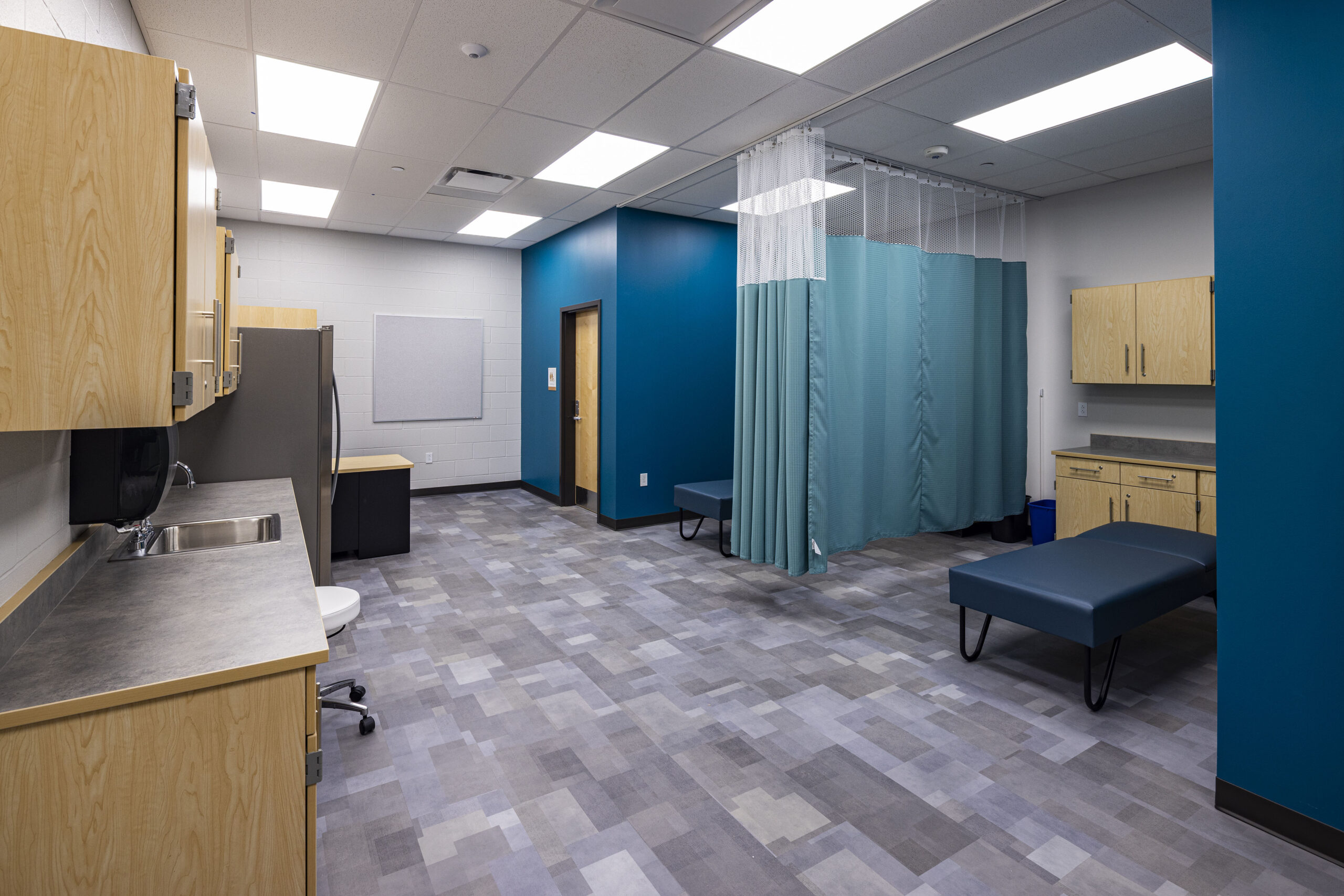 Medical examination room with blue walls, gray flooring, wooden cabinets, two examination tables, sinks, a desk, and a privacy curtain dividing the space. The room is clean, modern, and well-lit with ceiling lights.