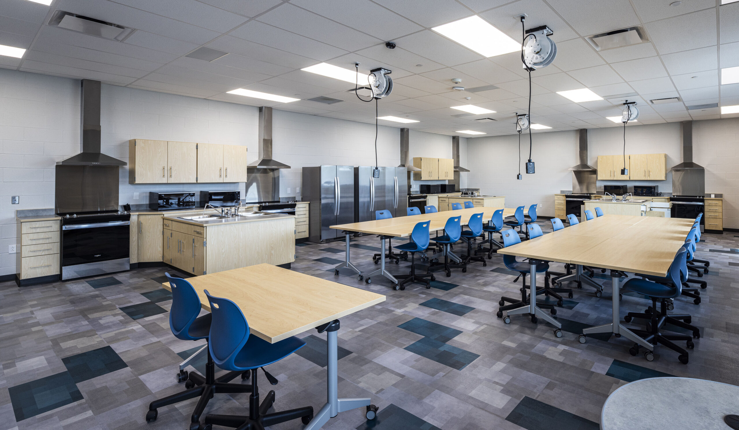 Modern classroom kitchen with multiple cooking stations, stainless steel appliances, wooden cabinets, and blue chairs around light wood tables. The room has a tiled floor and ceiling-mounted electrical outlets.