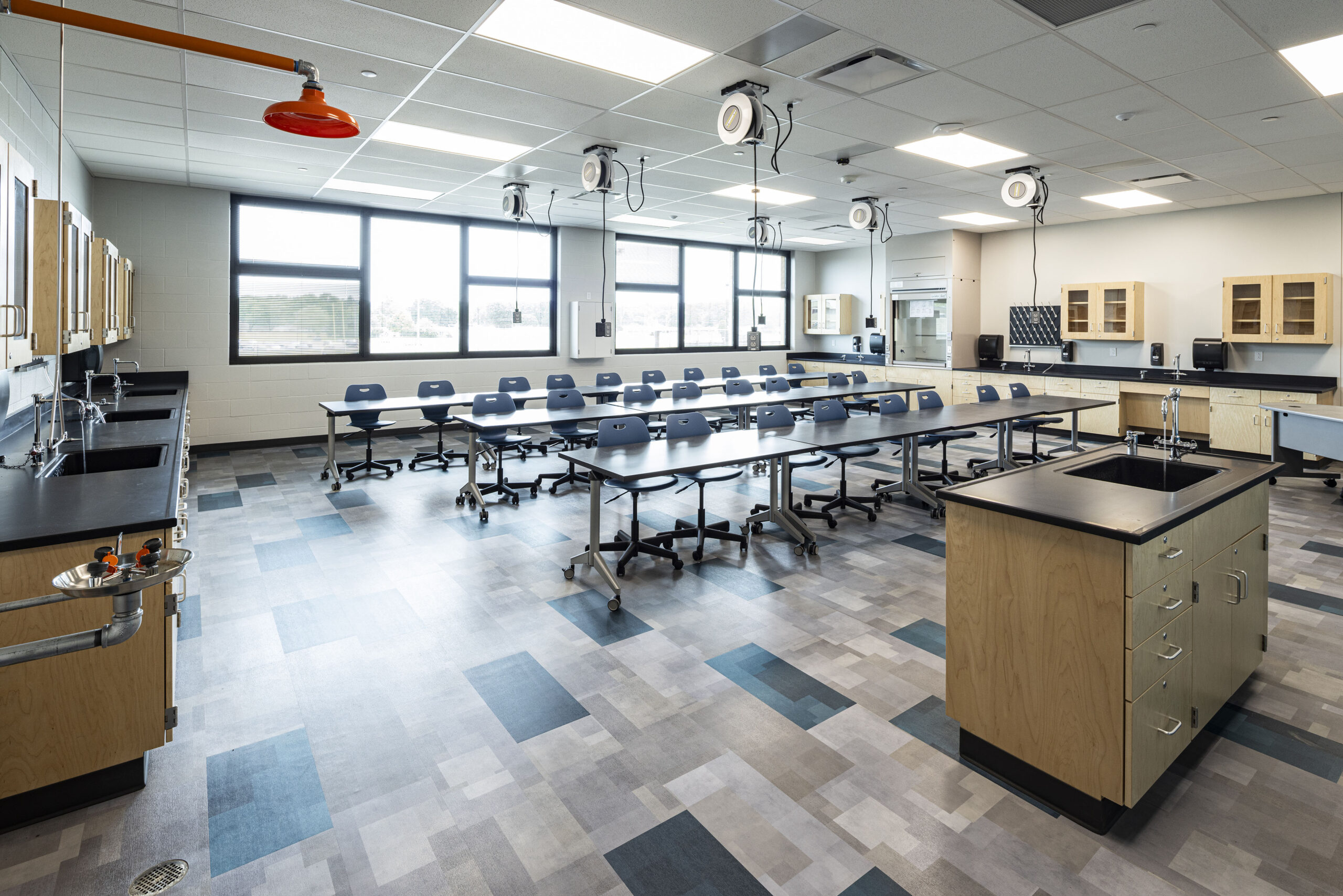 A modern science classroom with lab tables, black chairs, sinks, cabinets, ceiling electrical reels, and large windows letting in natural light. The room is empty and brightly lit.