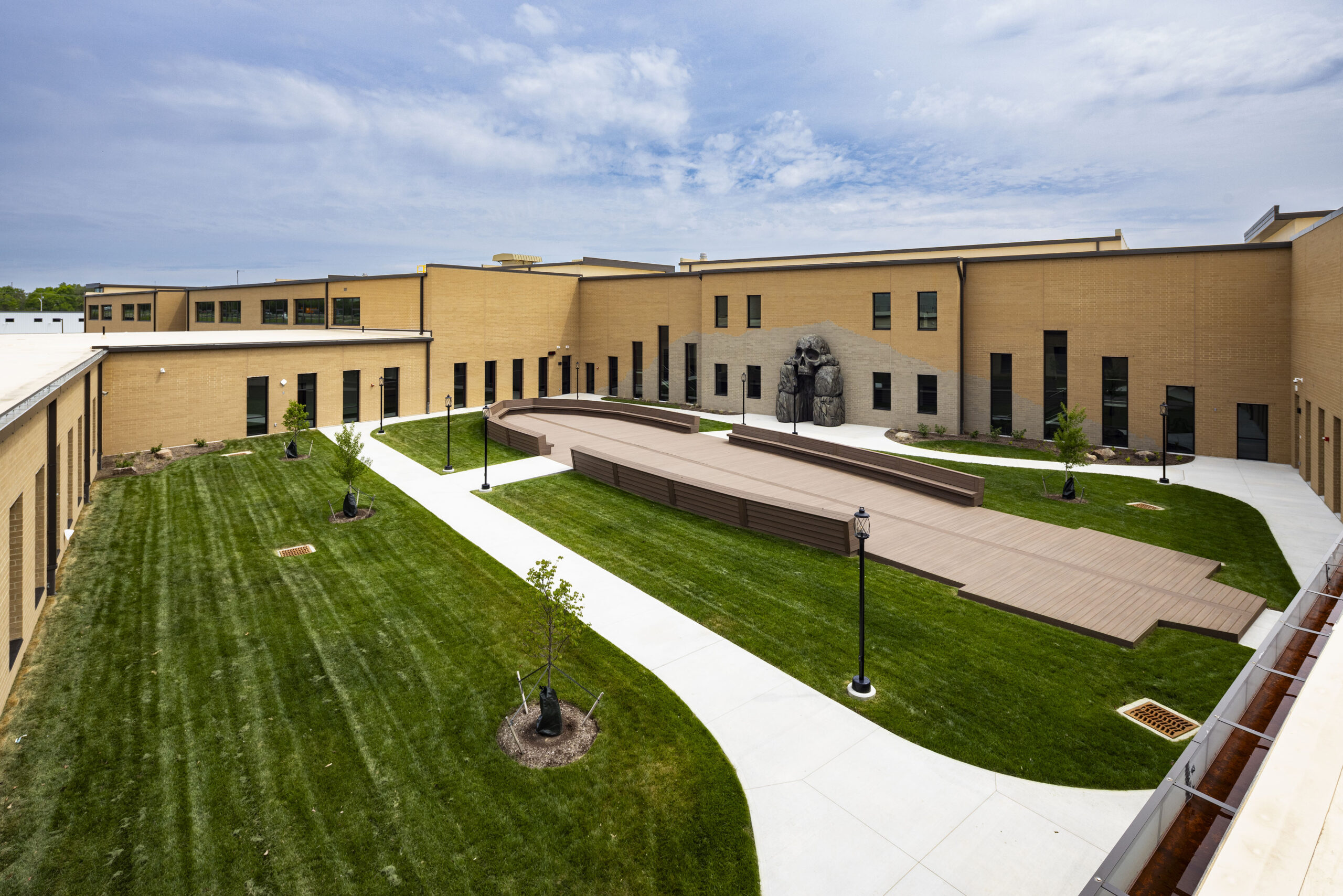 A courtyard with green grass, sidewalks, young trees, a wooden deck area, and a sculpture against a tan brick building under a partly cloudy sky.