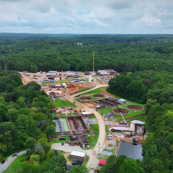 Aerial view of a large water treatment plant under construction, surrounded by dense green forest, with visible circular and rectangular tanks, roads, vehicles, and construction equipment.