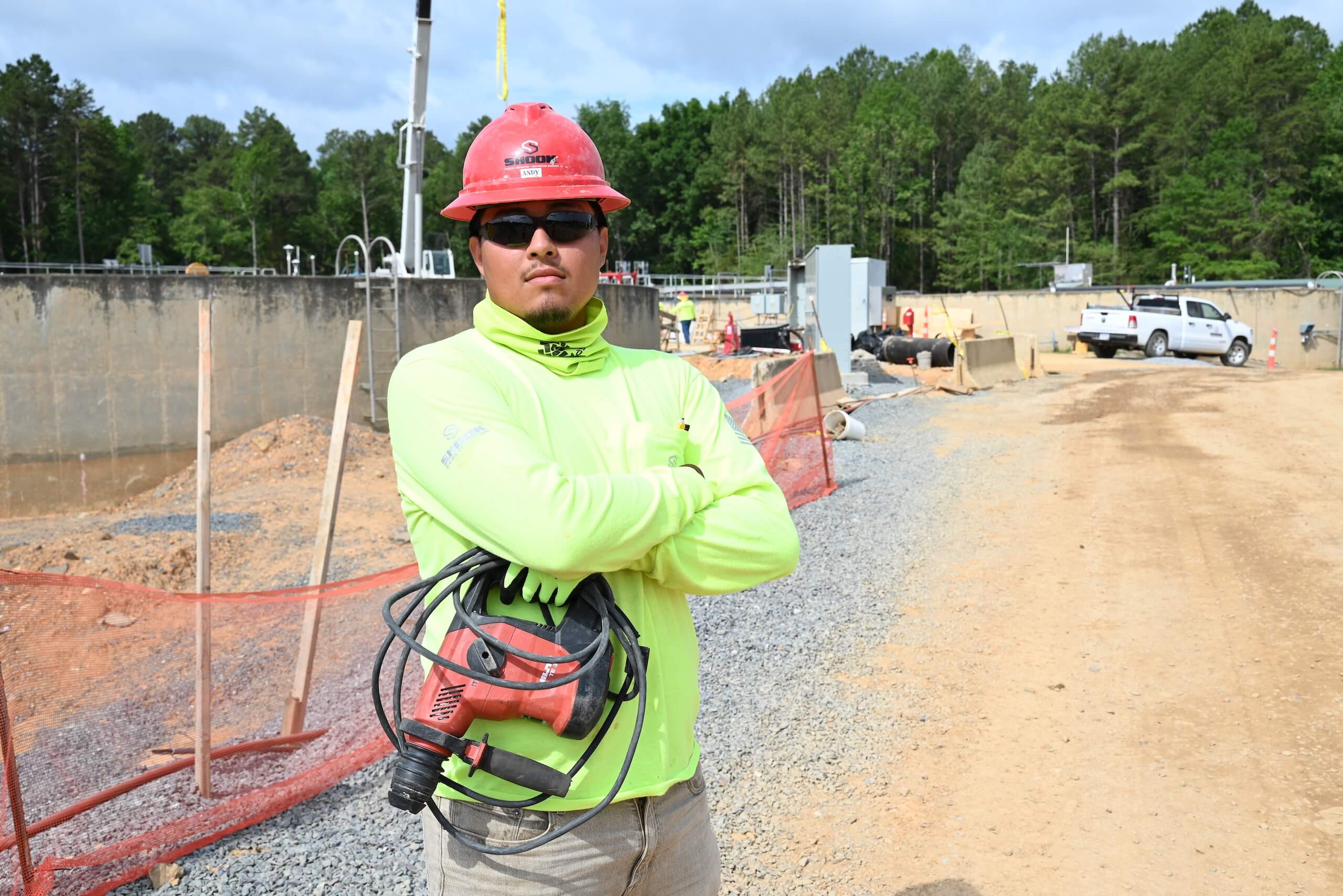 Construction worker in a neon shirt and red hard hat stands with arms crossed, holding a power tool, at a job site with gravel, fencing, and construction equipment in the background.