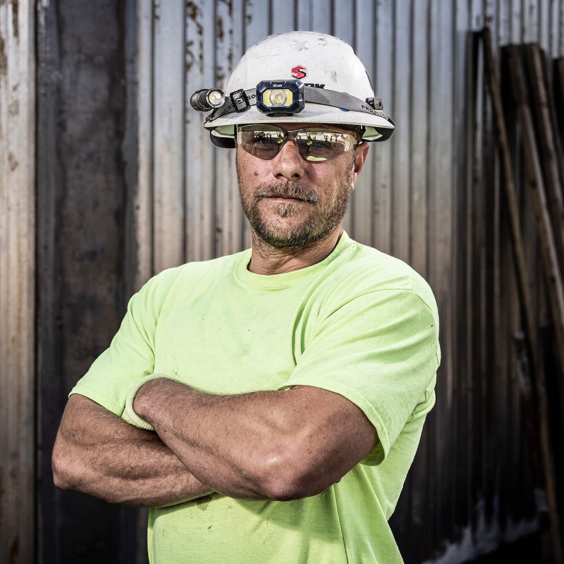 A construction worker wearing a hard hat, safety glasses, and a neon yellow shirt stands with arms crossed in front of a corrugated metal wall.