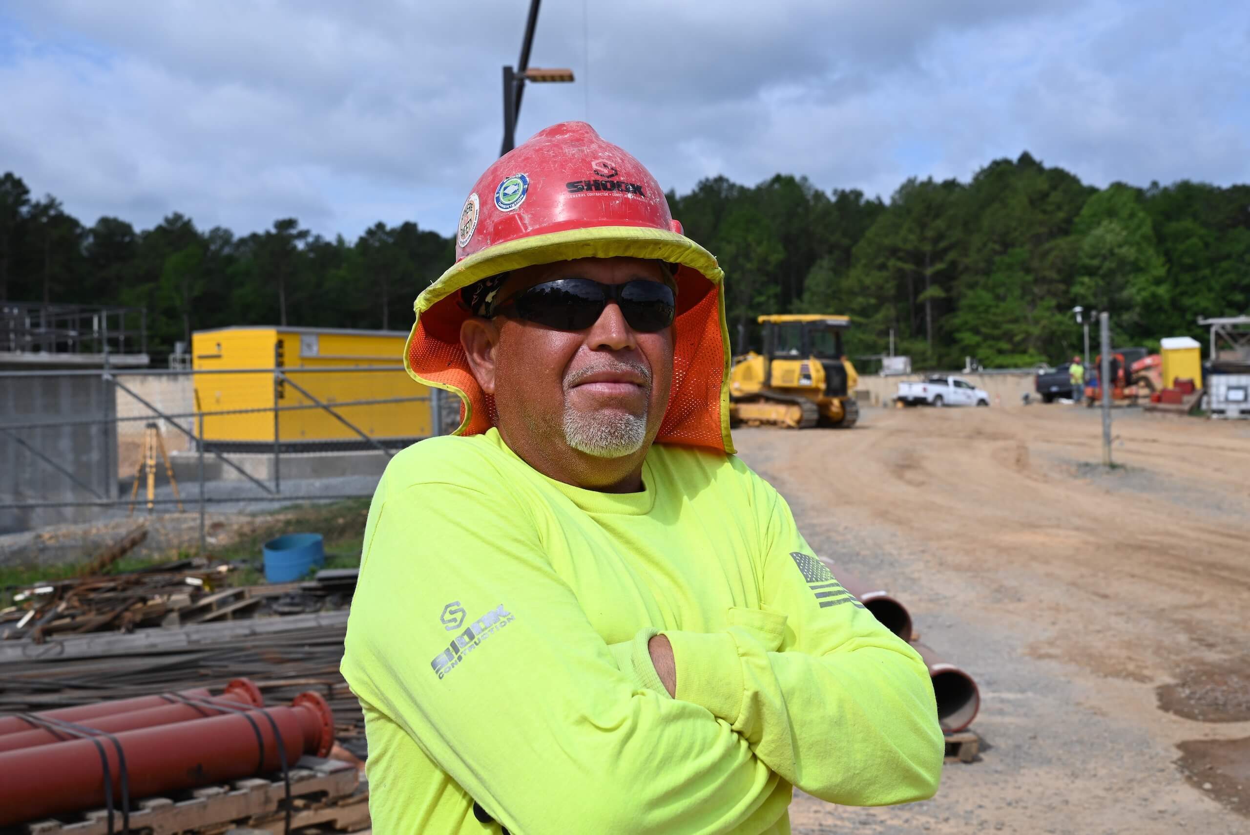 A construction worker in a bright yellow shirt and red hard hat stands with arms crossed at a worksite, wearing sunglasses. Construction equipment and vehicles are visible in the background.