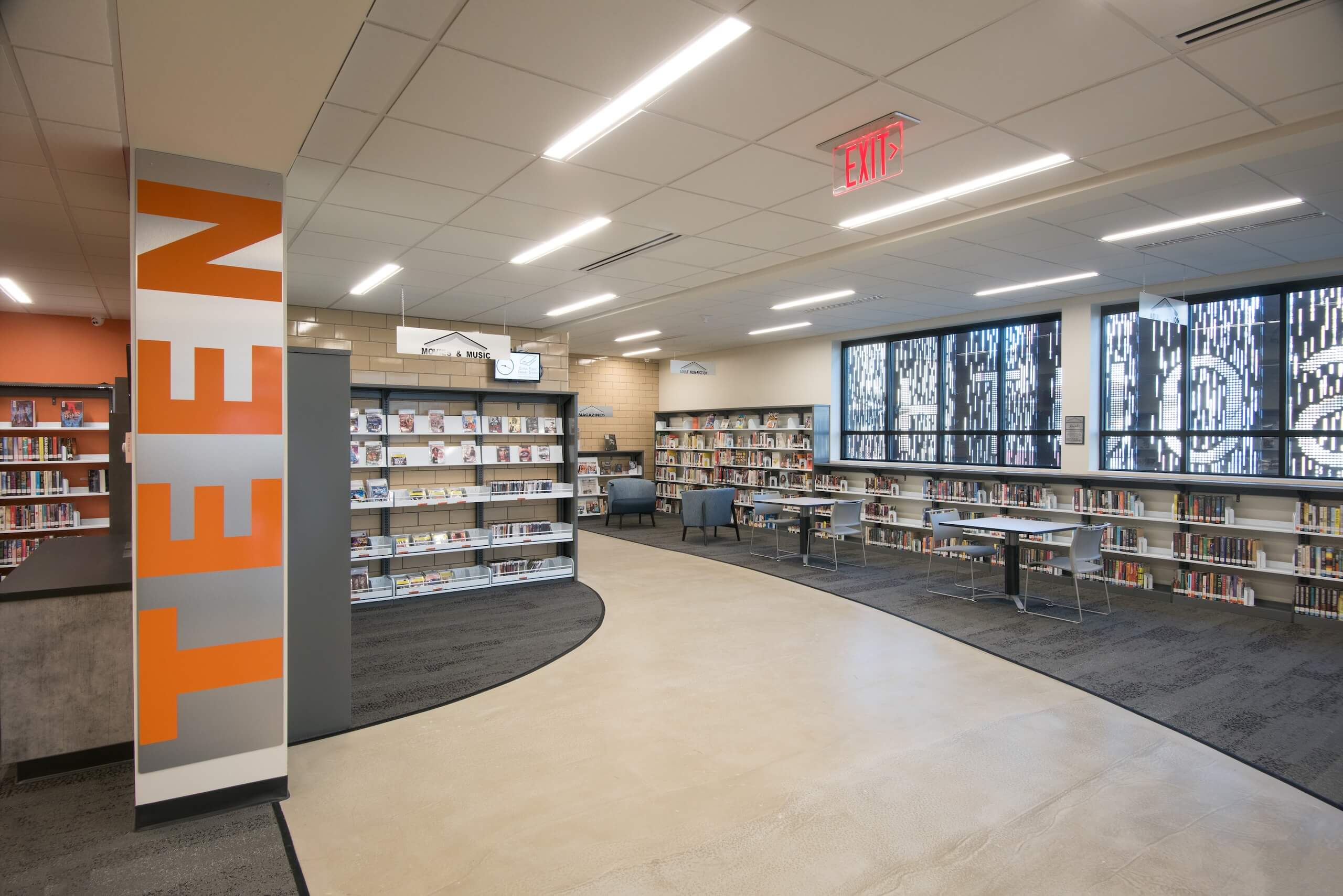 A modern library teen section with bookshelves, tables, chairs, and large windows. The word TEEN is printed in orange on a wall by the entrance. The space is bright and organized.