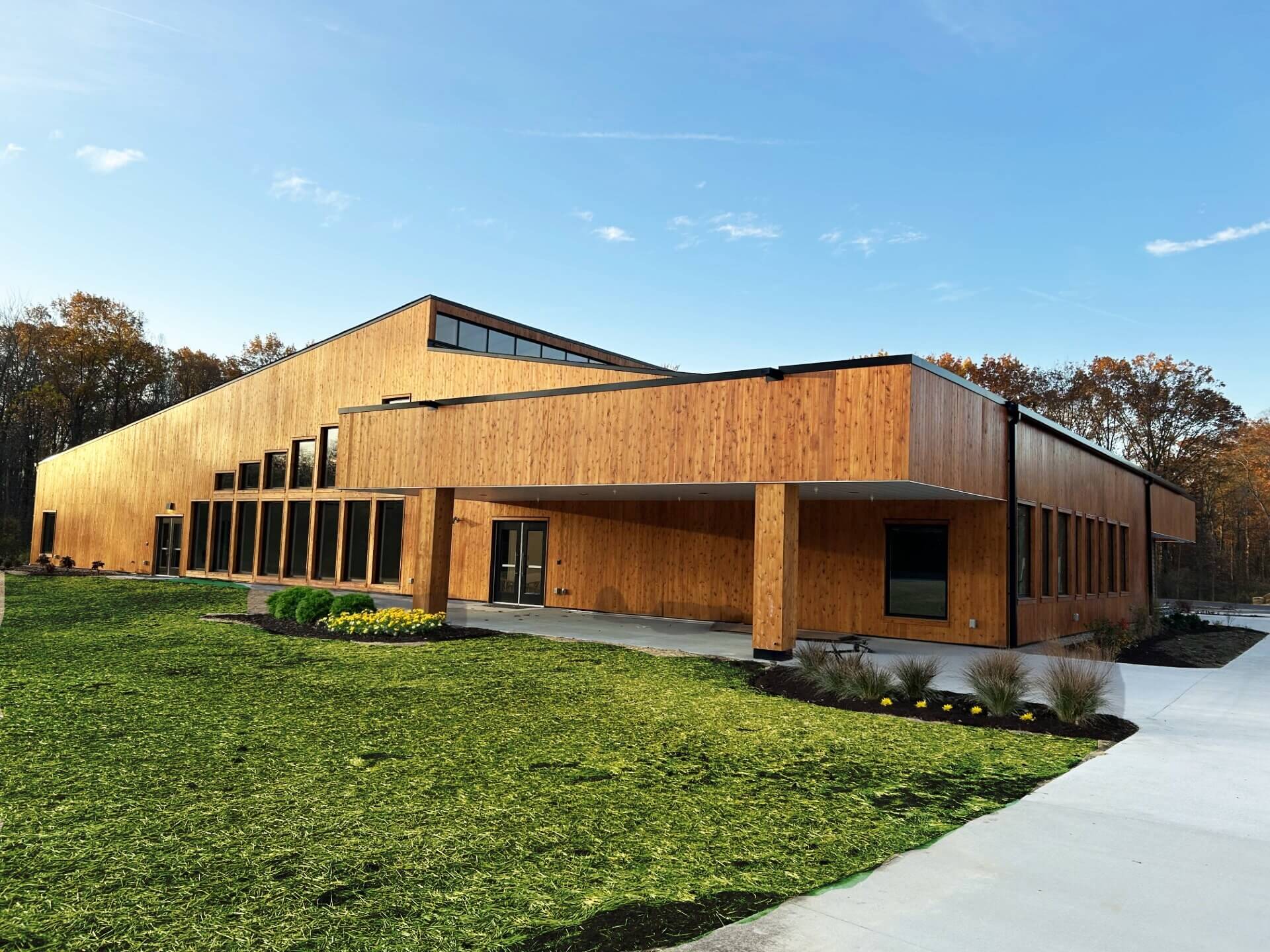 A modern, angular building with wood paneling, large windows, and a covered entrance sits beside a sidewalk and freshly landscaped grass under a blue sky.