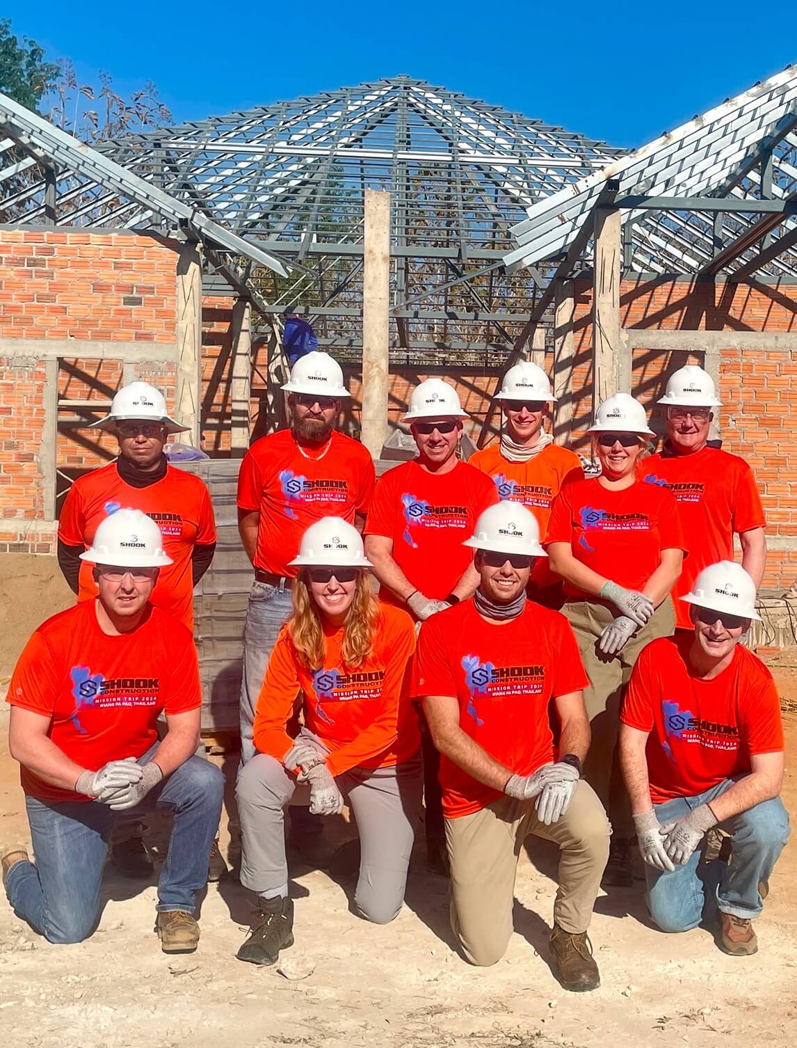 Ten people in orange shirts and white hard hats kneel and stand in front of a partially built brick structure with a steel roof frame under a clear blue sky, posing for a group photo at a construction site.