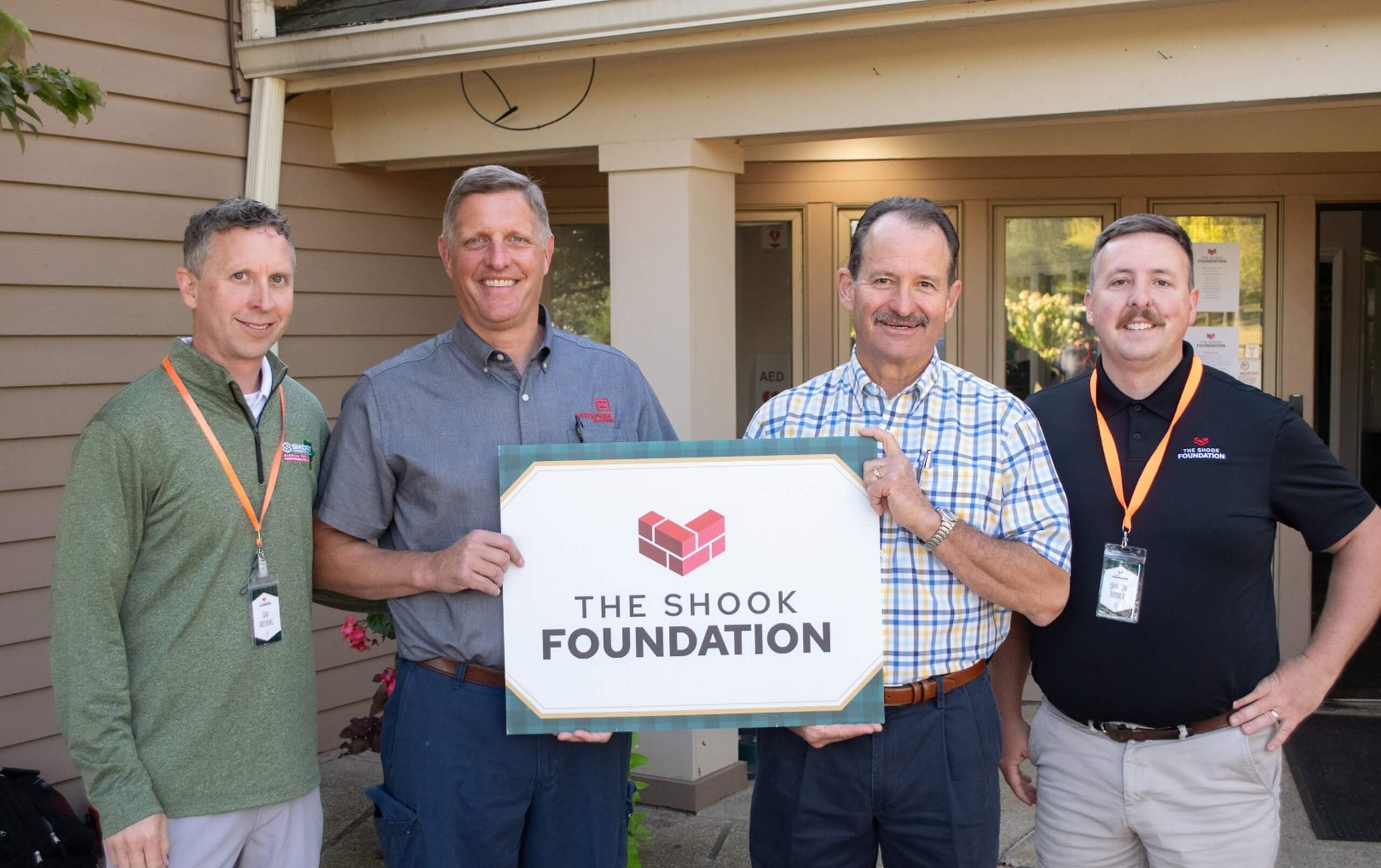 Four men stand outside a building, smiling, with two of them holding a large sign that reads The Shook Foundation. They are all wearing name badges and casual business attire.
