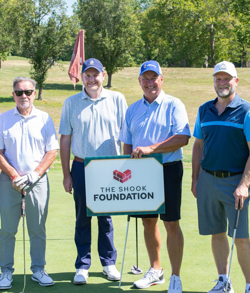 Four men stand on a golf course holding clubs, smiling next to a sign that reads The Shook Foundation. Trees and grass are visible in the background on a sunny day.