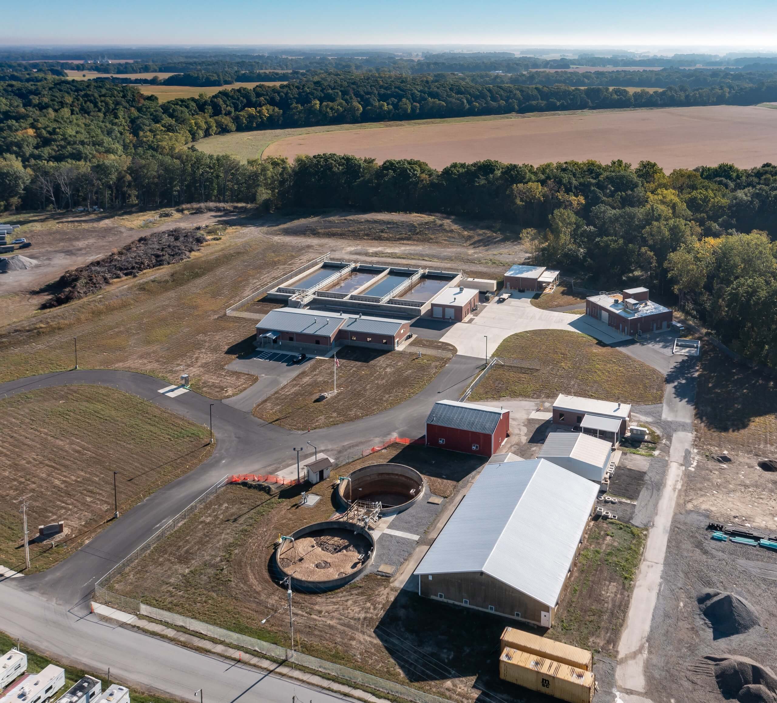 Aerial view of a water treatment facility surrounded by fields and trees, with several buildings, tanks, and paved roads throughout the property under a clear sky.