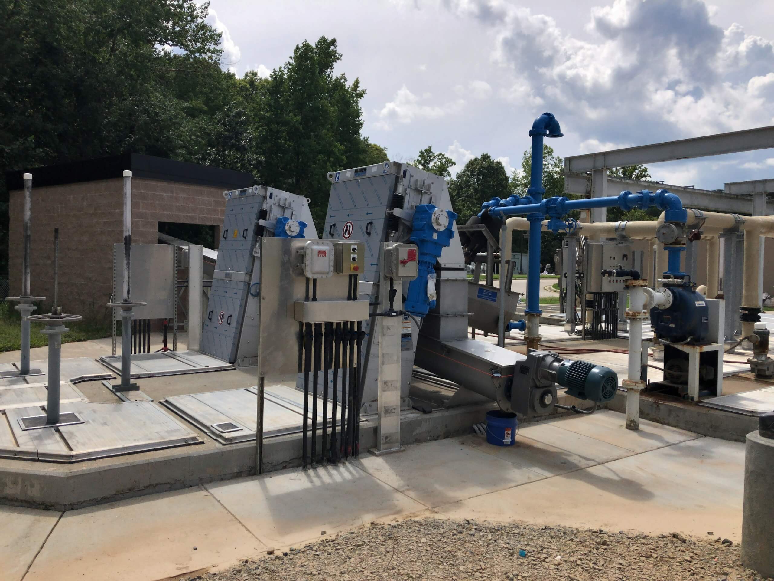Outdoor water treatment facility with various pipes, pumps, valves, and metal equipment. Trees and a small building are in the background under a partly cloudy sky.