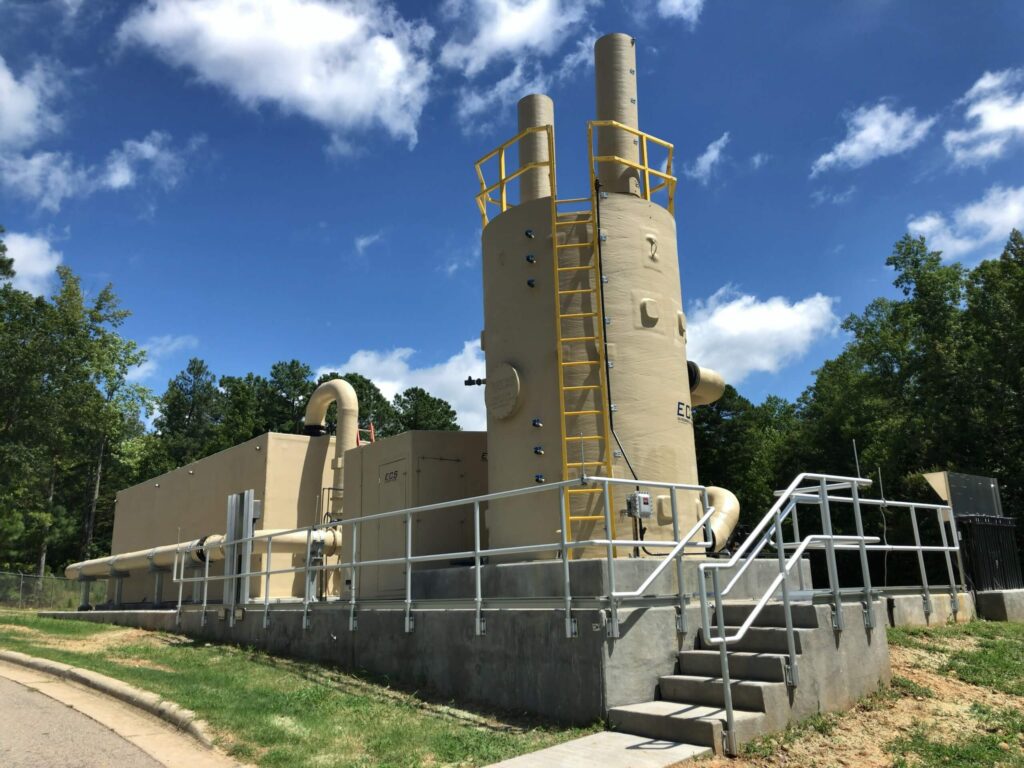 Large industrial scrubber system with tall cylindrical towers, yellow safety railings, and metal stairs, located outdoors on a concrete platform, surrounded by trees under a partly cloudy blue sky.