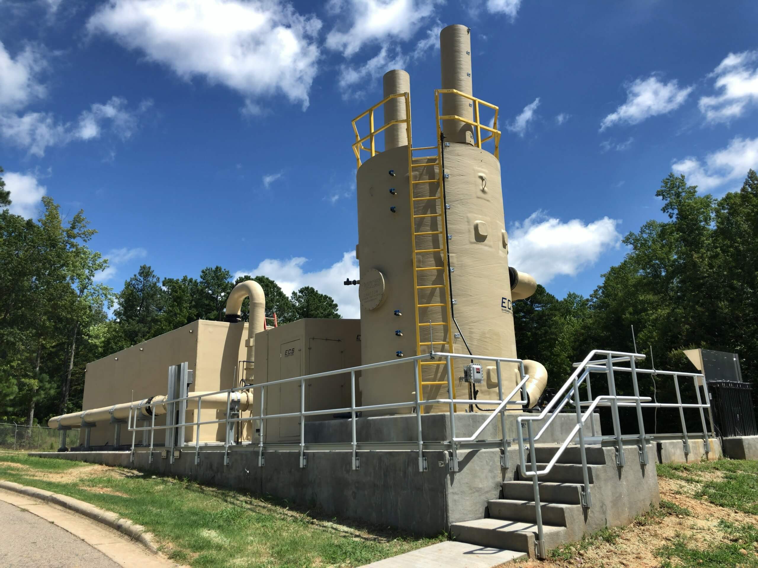 Large industrial scrubber system with tall cylindrical towers, yellow safety railings, and metal stairs, located outdoors on a concrete platform, surrounded by trees under a partly cloudy blue sky.