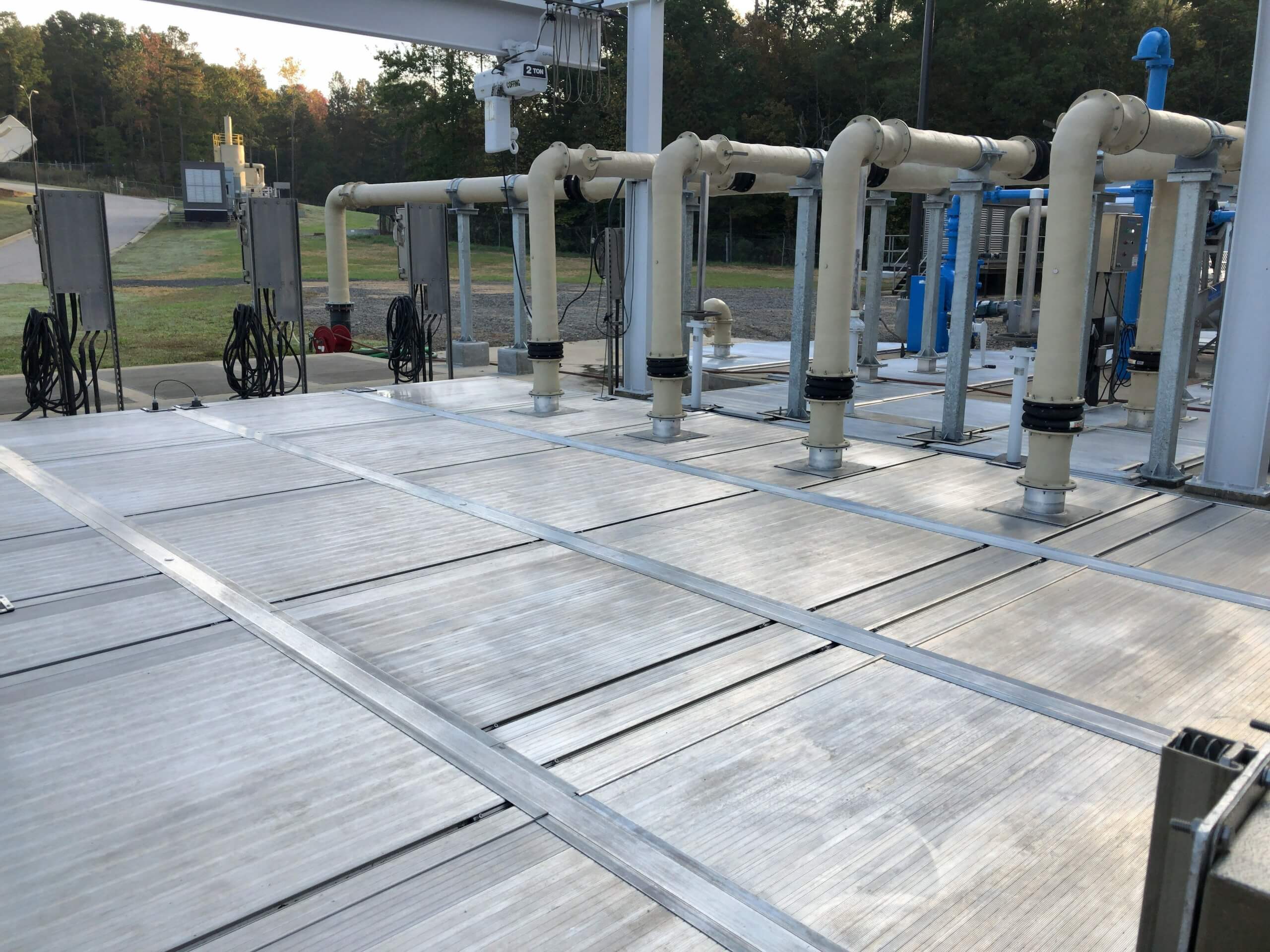 Industrial site with rows of large white pipes and metal grates on the ground, surrounded by trees and outdoor equipment, under a clear sky.