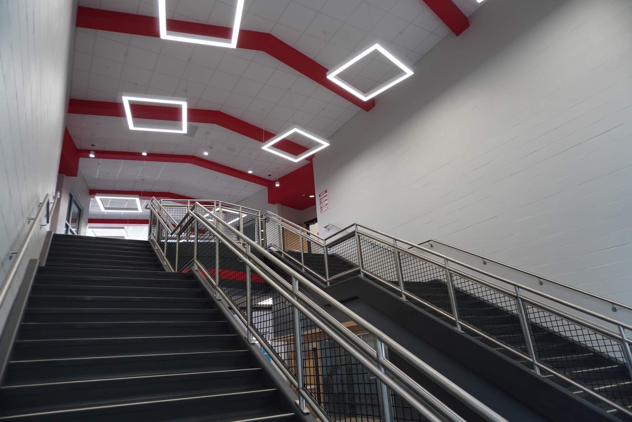 Modern indoor staircase with black steps, silver railings, and geometric ceiling lights. Red beams and white walls create a clean, contemporary look. Multiple stair flights lead up to another floor.