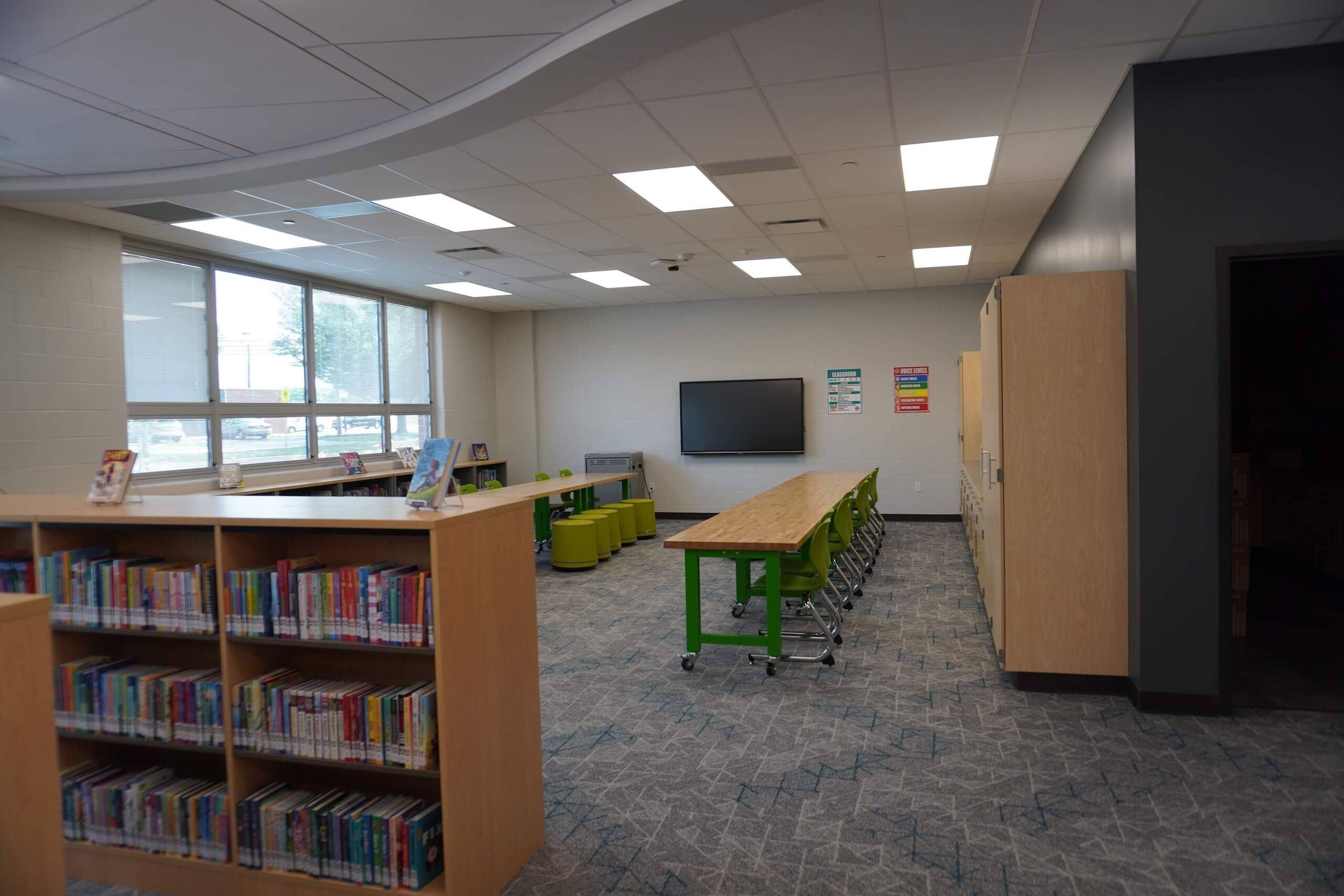 A modern library room with bookshelves, a long table with green chairs, a wall-mounted screen, and posters on the wall. Large windows let in natural light.