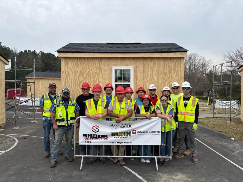A group of construction workers and students in safety gear stand in front of two wooden sheds, holding a banner for a high school construction project. Scaffolding is visible beside the sheds.