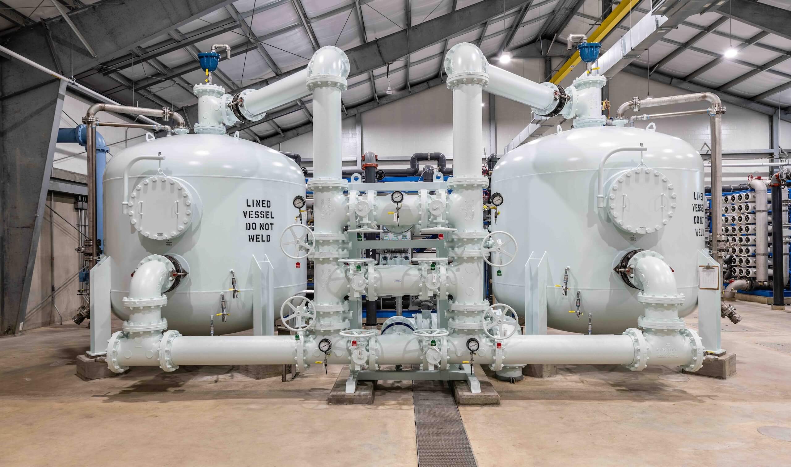 Two large, white industrial pressure vessels connected by multiple pipes and valves inside a spacious facility. One vessel is labeled LINED VESSEL DO NOT WELD. The area is well-lit with exposed ceiling beams.