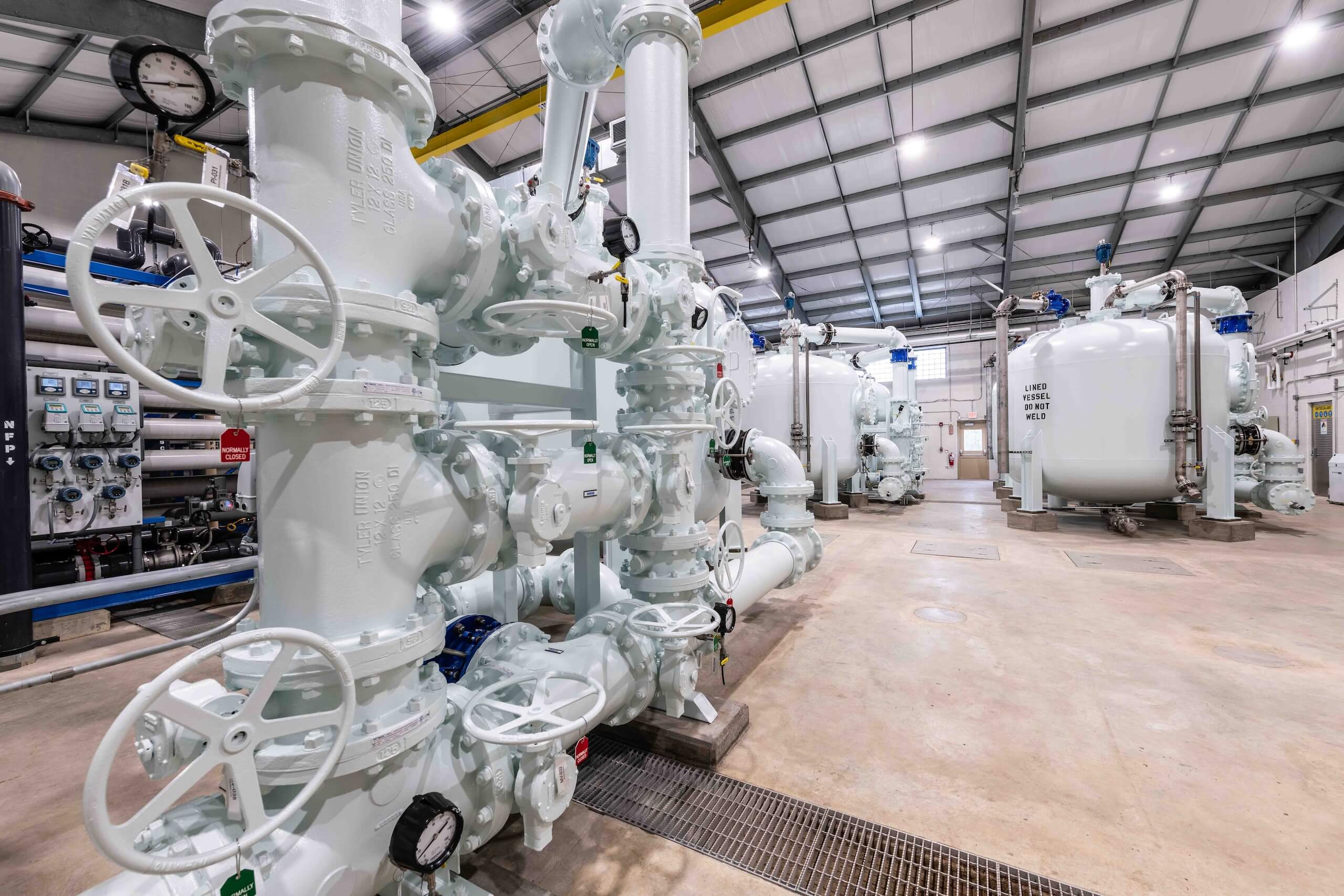 Large industrial room with metal pipes, valves, gauges, and tanks painted white. The space has a high ceiling with exposed beams and a concrete floor. Equipment appears organized and well-maintained.