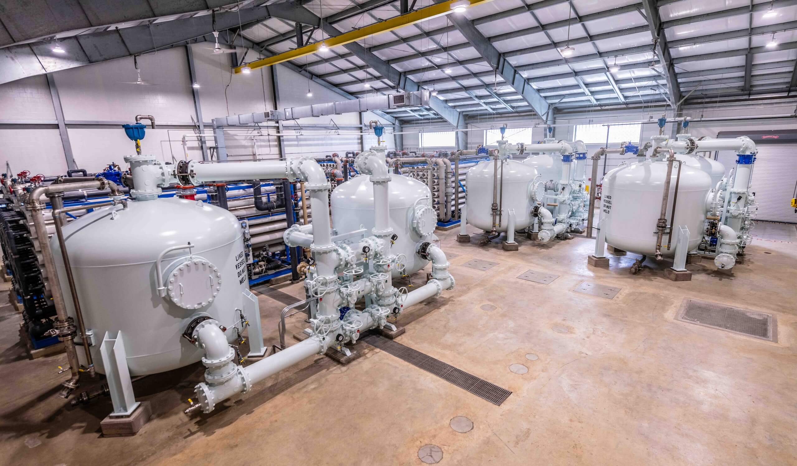 Large industrial room with four white cylindrical tanks connected by pipes, valves, and gauges, under a high metal roof with skylights. The floor is clean, and the tanks are evenly spaced in the spacious facility.