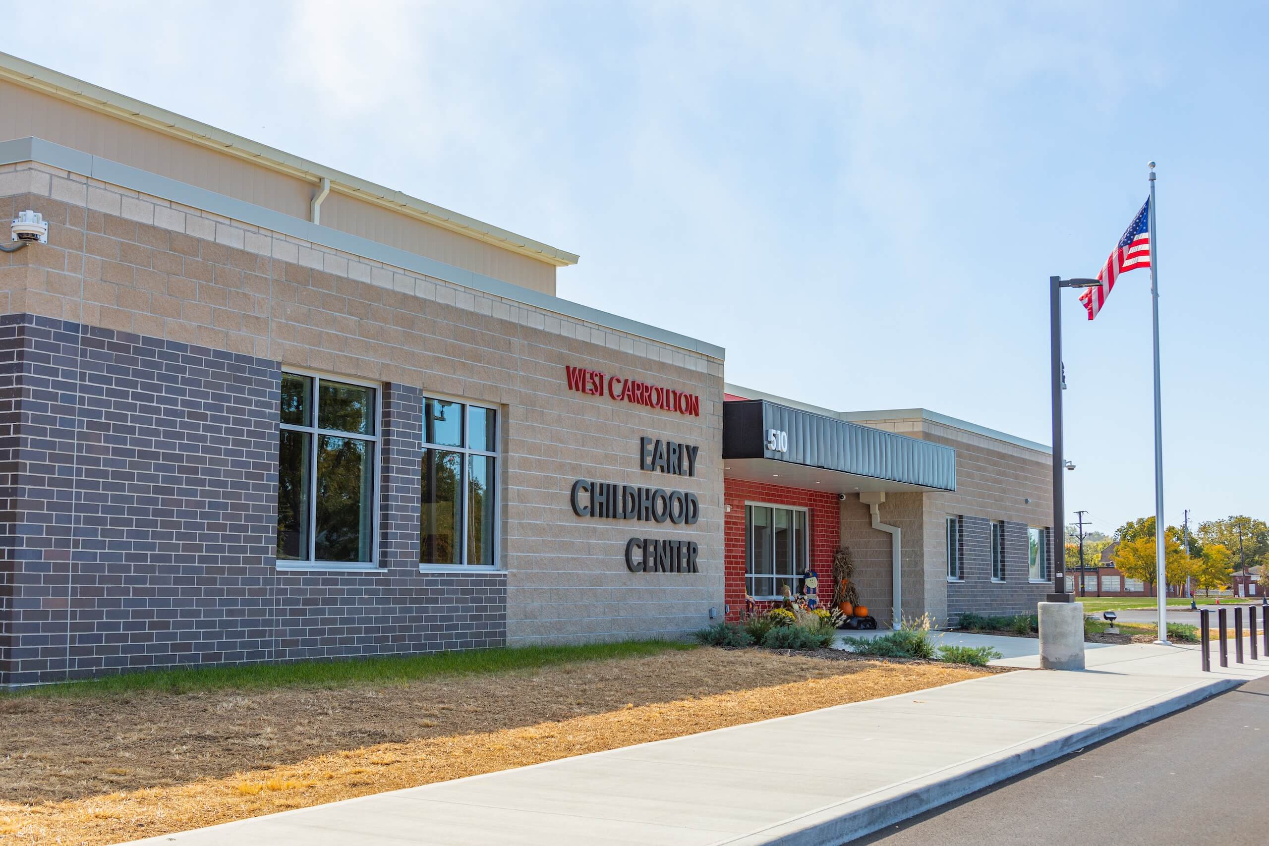 Modern school building with a sign reading West Carrolton Early Childhood Center, large windows, a sidewalk, landscaping, and a flagpole flying the U.S. flag in front. The sky is clear and sunny.