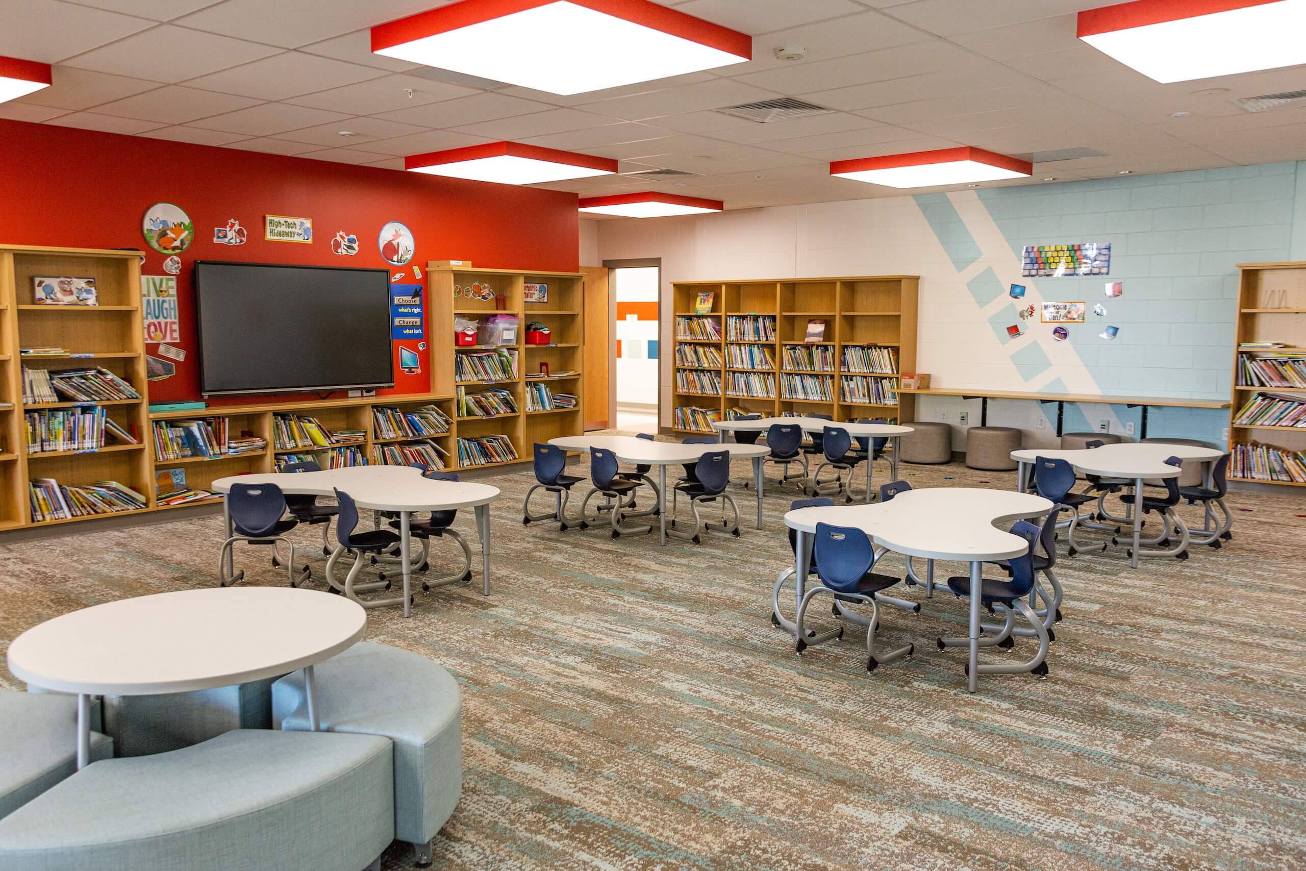 Modern library with round tables, blue chairs, bookshelves along the walls, and a large screen at the front. The room is brightly lit with a red and white ceiling design and colorful decor.