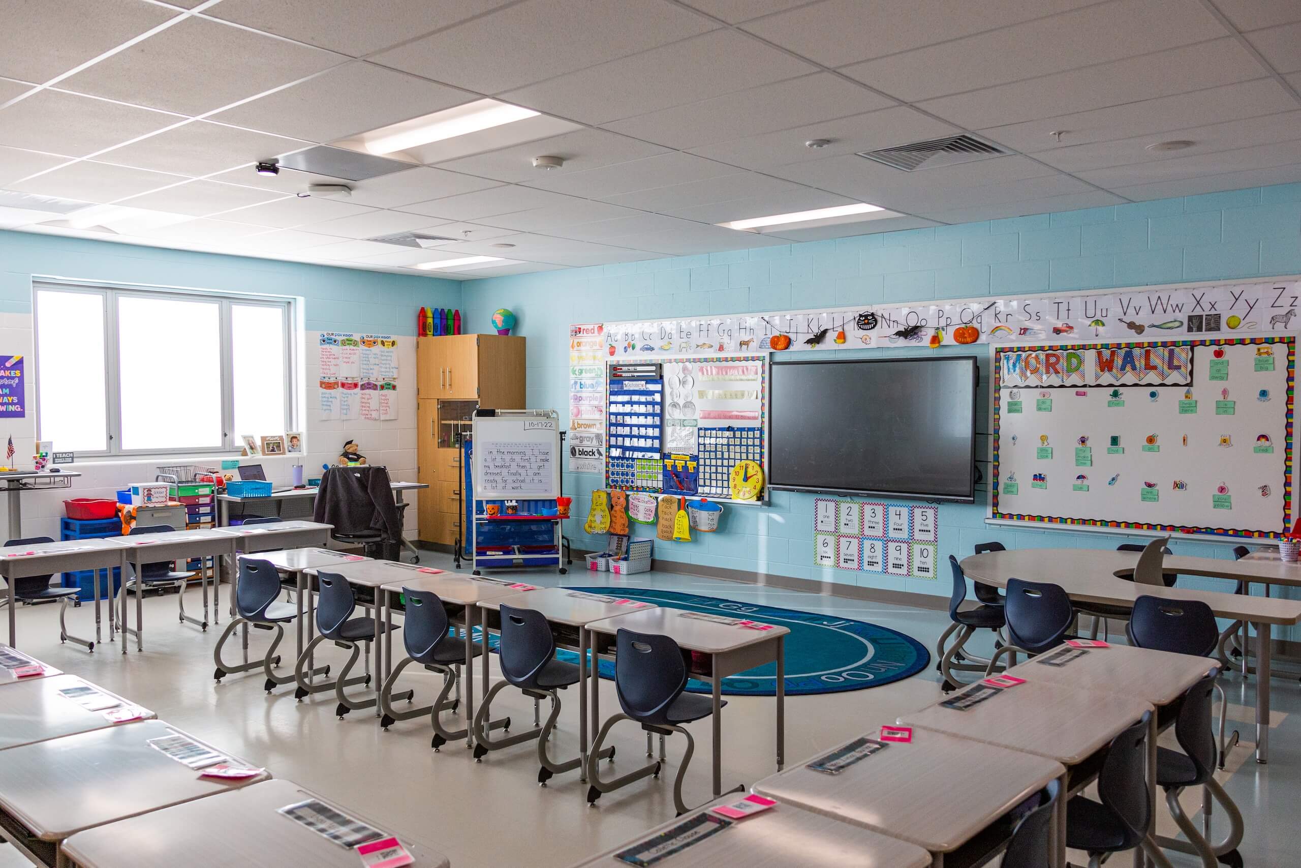 A bright, empty classroom with desks in rows, blue chairs, colorful decorations, a large digital display, whiteboards, learning charts, and educational materials neatly arranged. Sunlight streams through large windows.