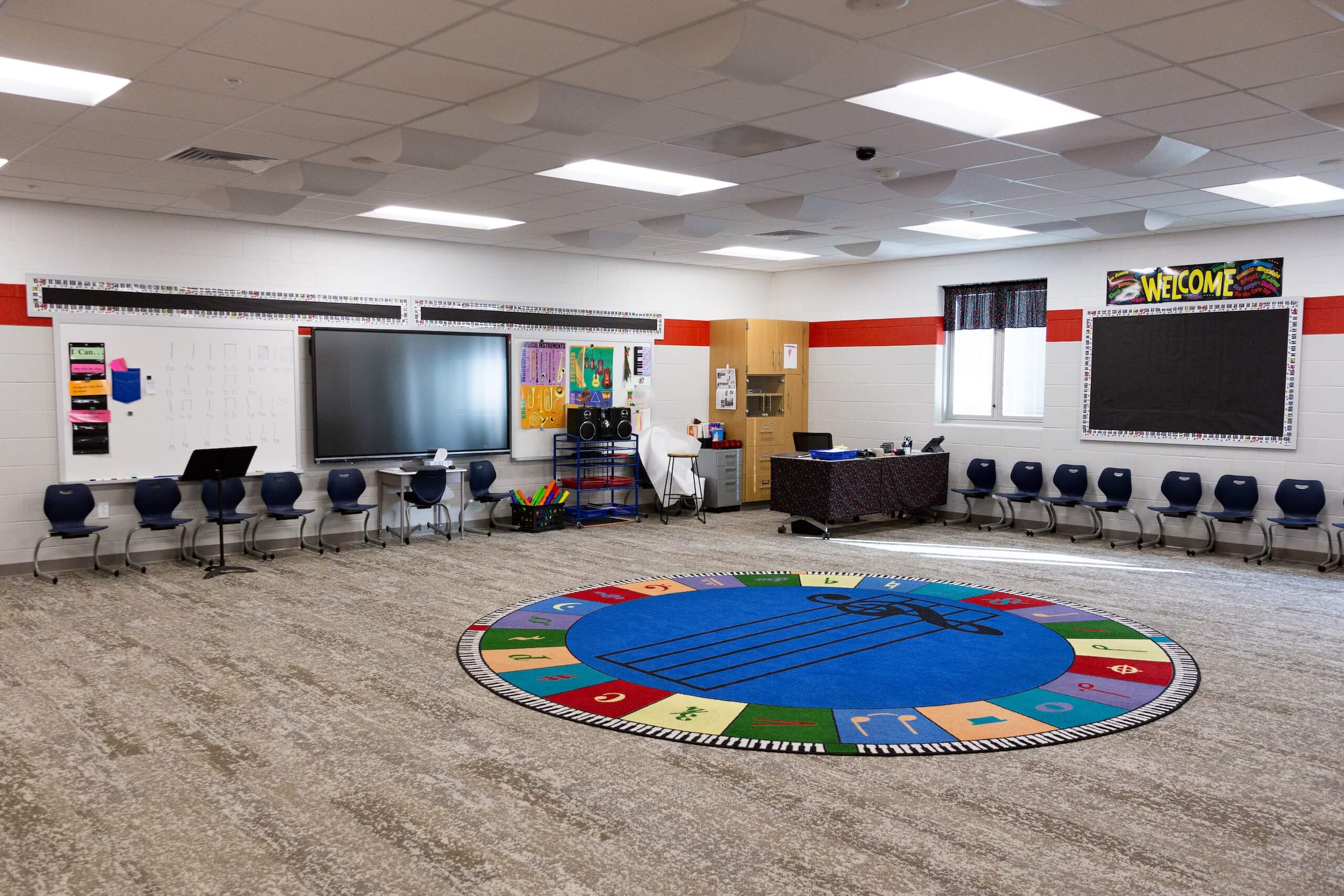 A spacious classroom with chairs along the walls, a large interactive screen, posters, and a blue circular rug with music notes in the center. The walls have a welcome sign and display boards.