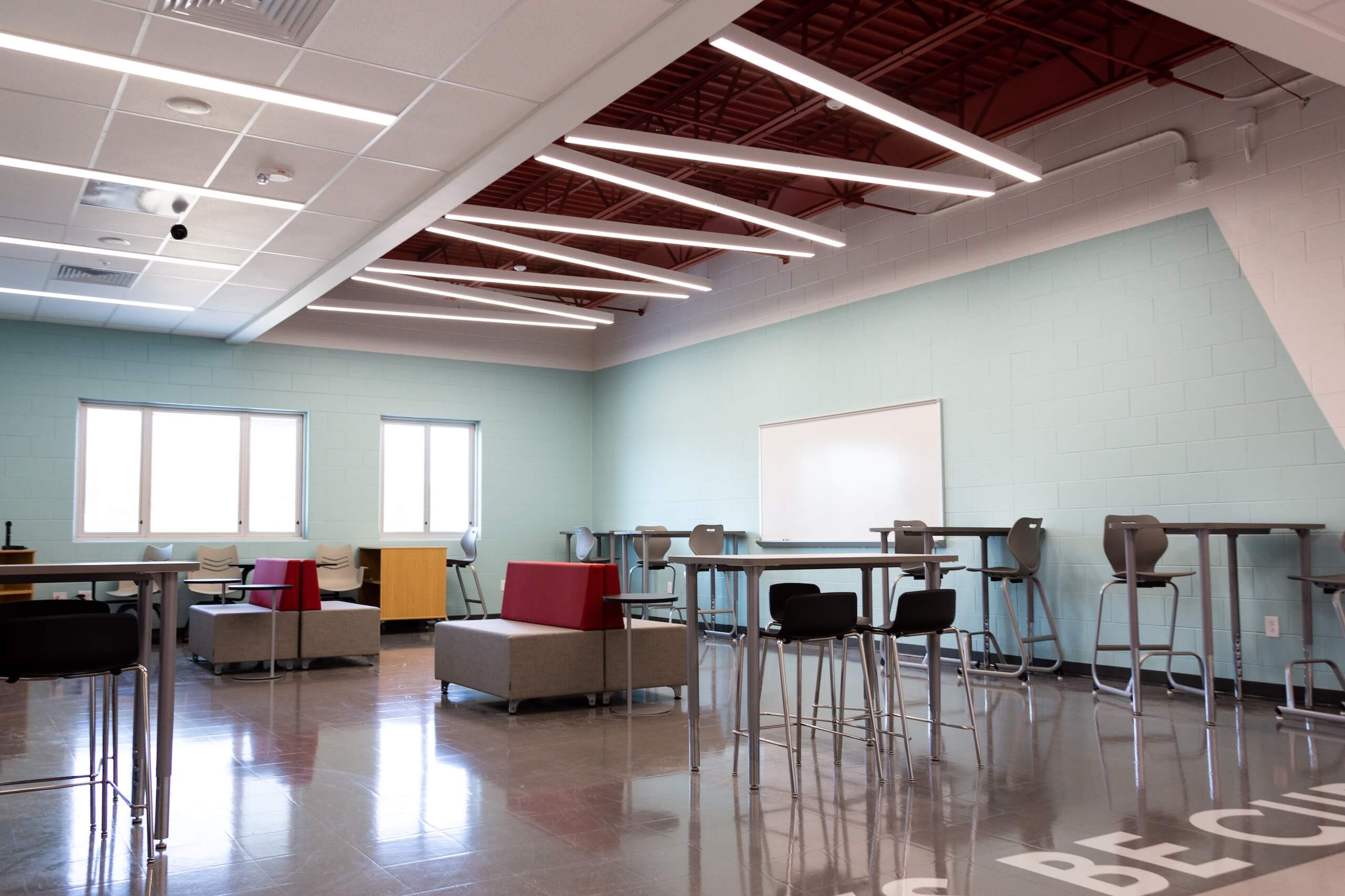 Modern classroom with high tables, stools, lounge seating, large windows, a whiteboard, and exposed ceiling beams. The room is bright, spacious, and has a polished floor.