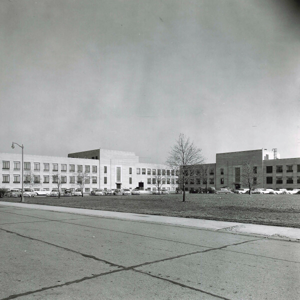 Black-and-white photo of a large, three-story institutional building with parked cars in front. A leafless tree and a street are visible in the foreground.