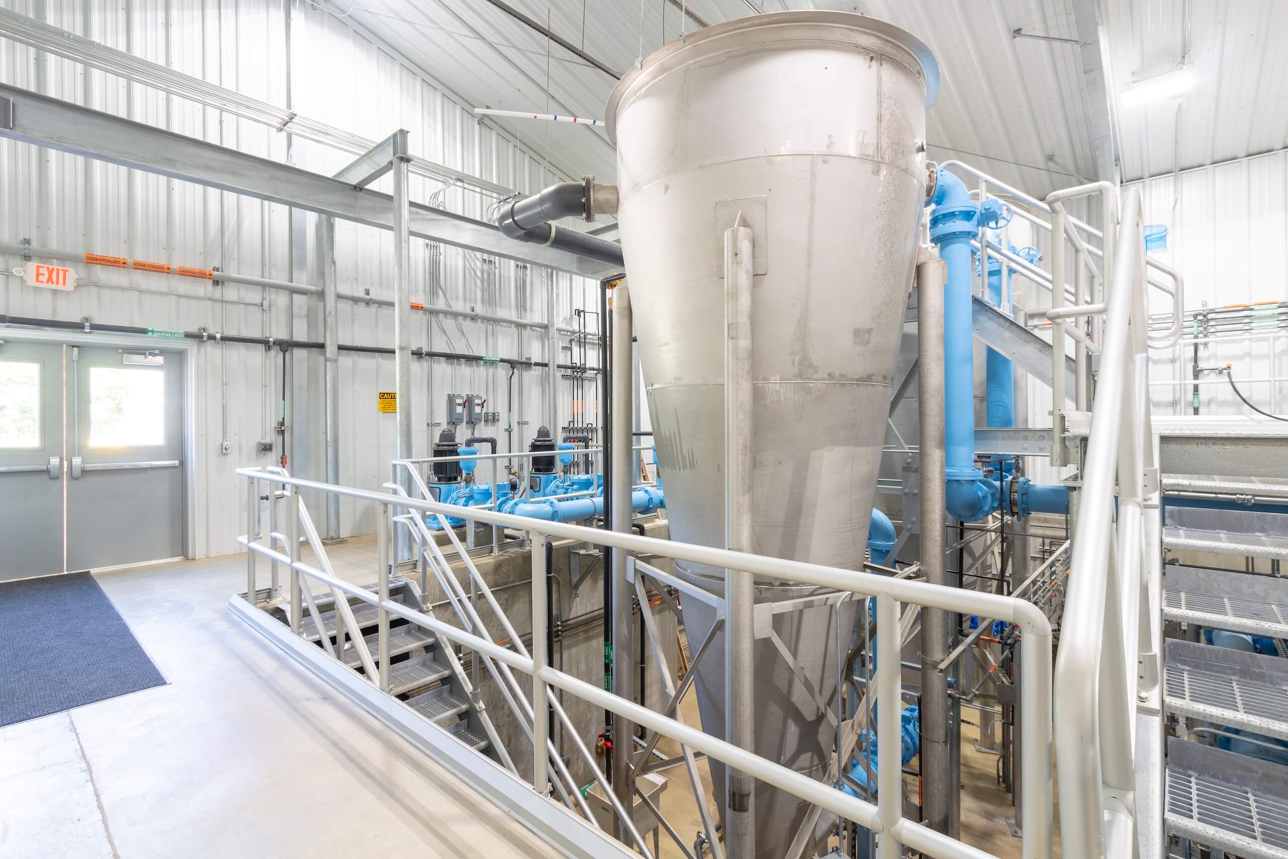 A large silver industrial tank with pipes and railings inside a bright, modern processing facility with metal walls and ceiling. Blue machinery and exit doors are visible in the background.