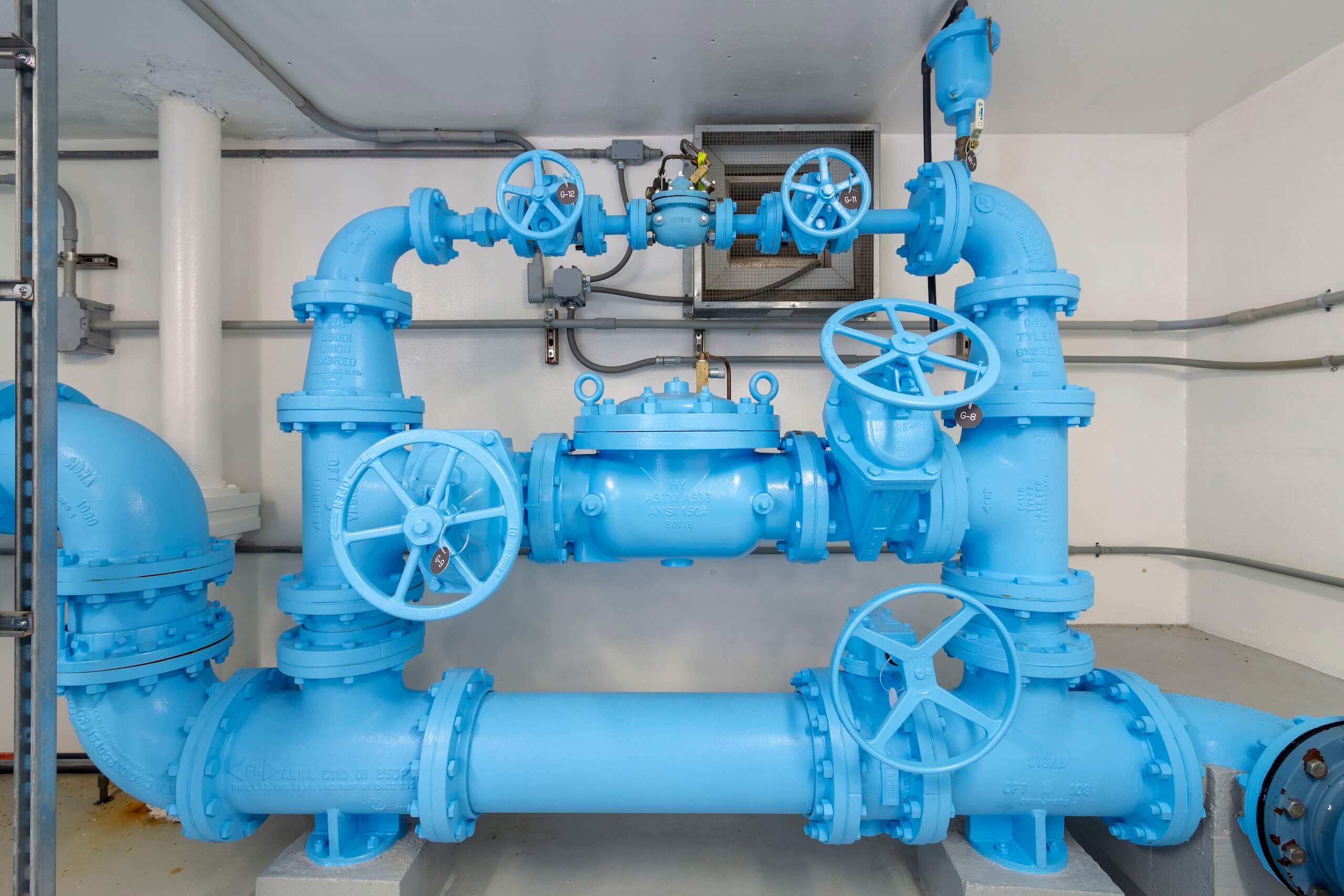 A complex system of blue industrial water pipes and valves mounted to a wall inside a utility room, with several large handwheels and connecting joints visible.