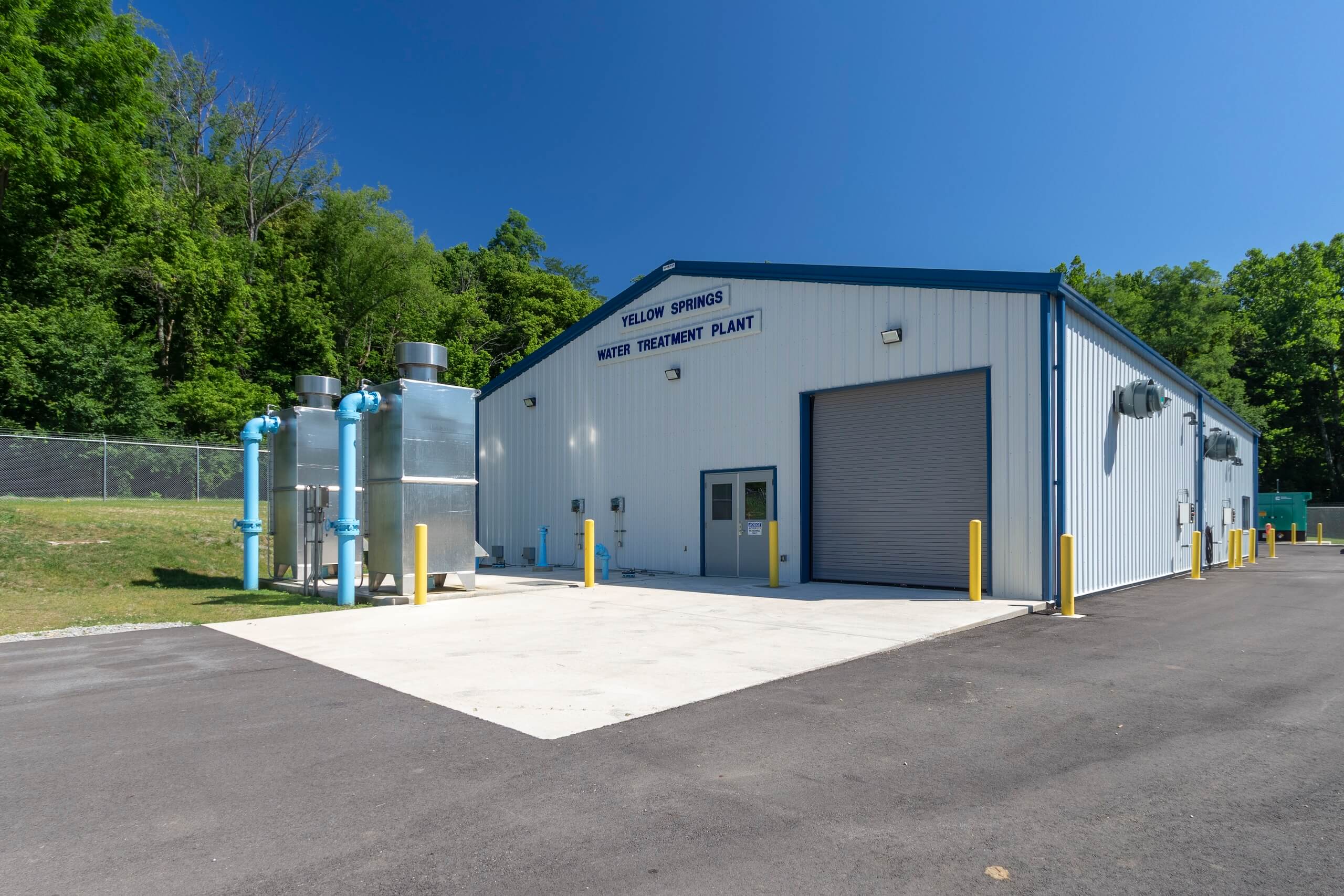 A small industrial building labeled Yellow Springs Water Treatment Plant with metal pipes and equipment outside, surrounded by trees and a clear blue sky.