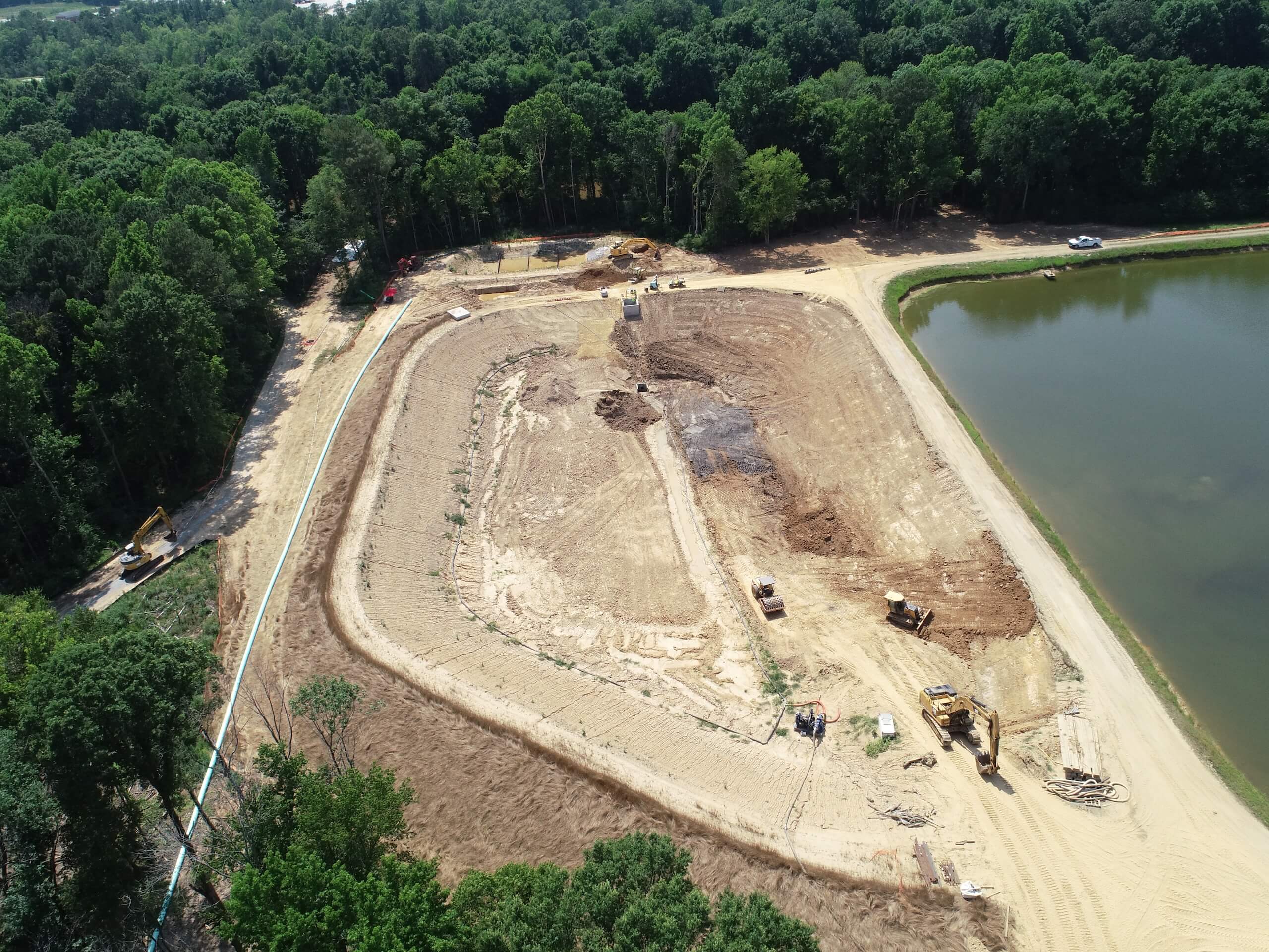 Aerial view of a construction site beside a pond, surrounded by trees. Dirt vehicles and construction materials are visible on the cleared land.