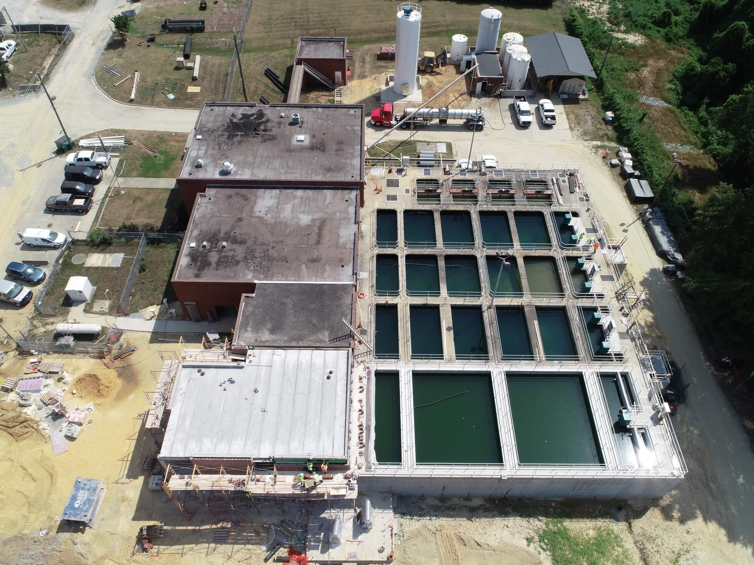 Aerial view of a water treatment facility with rectangular open tanks, adjacent buildings, storage tanks, vehicles, and construction materials on-site.