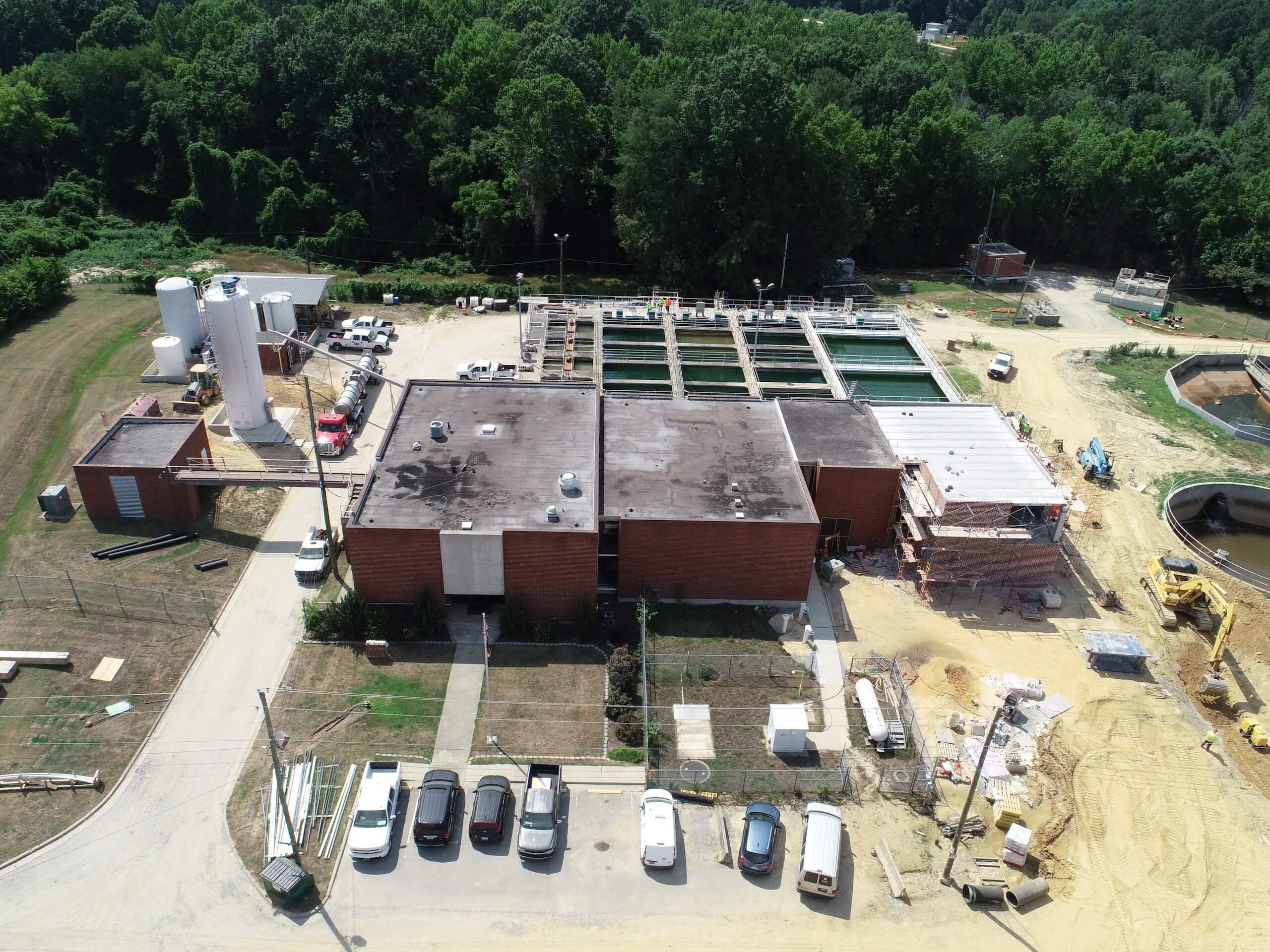 Aerial view of a water treatment facility with several tanks, brick buildings, parked cars, construction materials, and wooded area in the background.