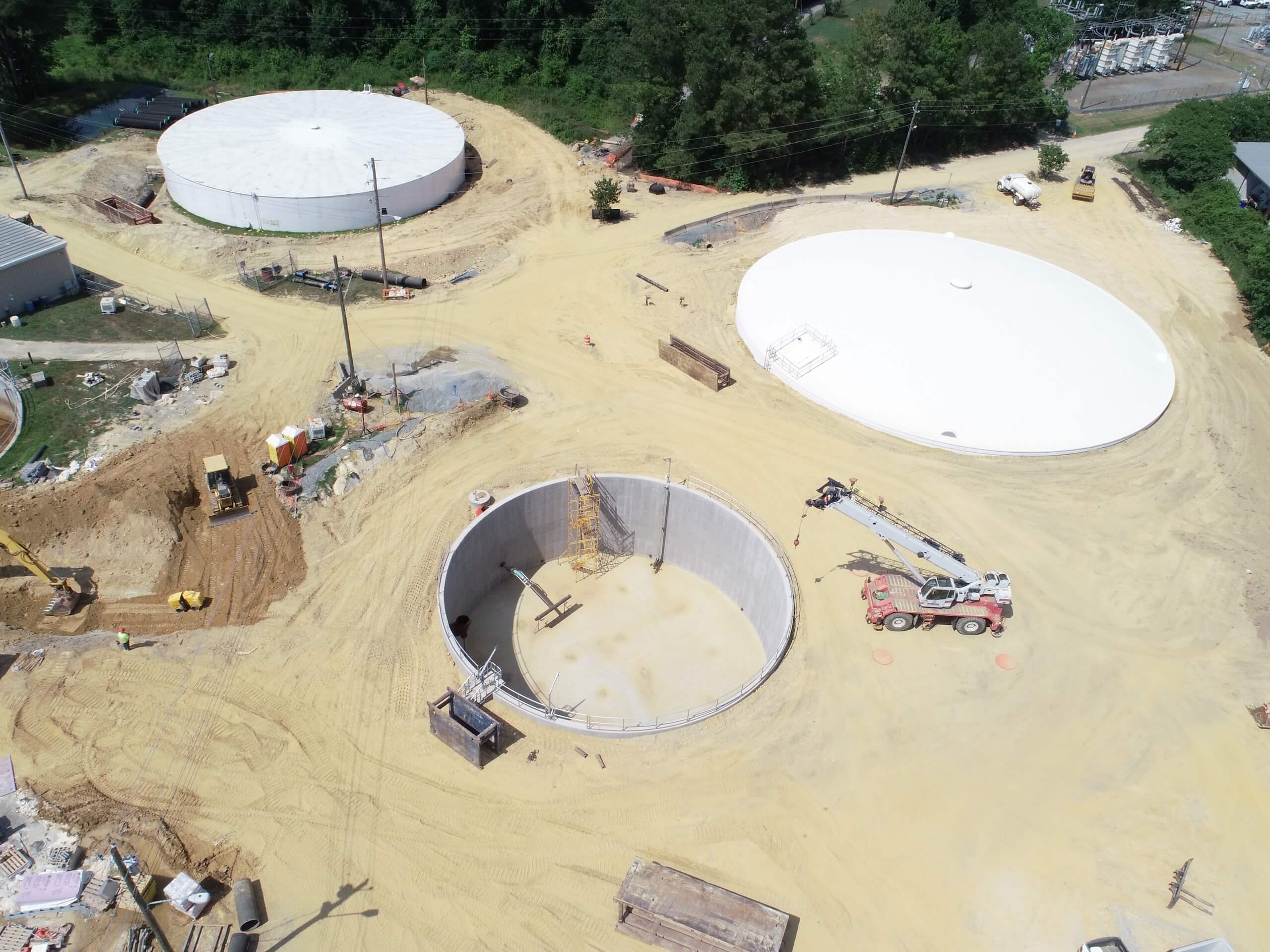 Aerial view of a construction site with three large circular tanks, sand-covered ground, construction equipment, and vehicles scattered around. Trees border the site in the background.