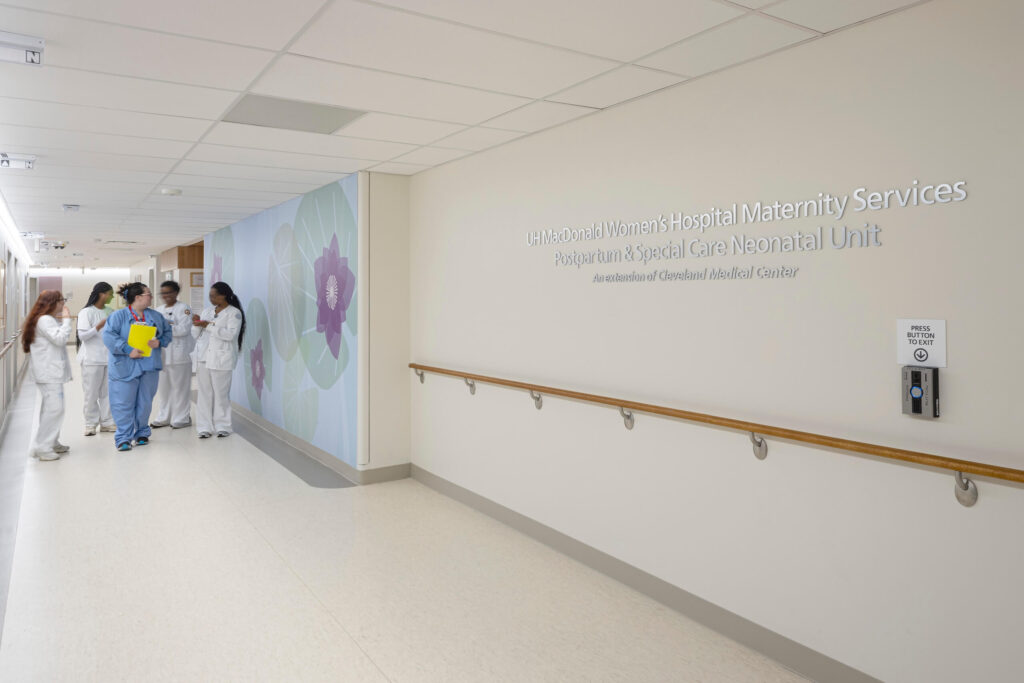 A group of healthcare workers in scrubs stand talking near a hospital corridor. A wall sign reads Maternity Services and Neonatal Unit next to a handrail and an accessible door button.