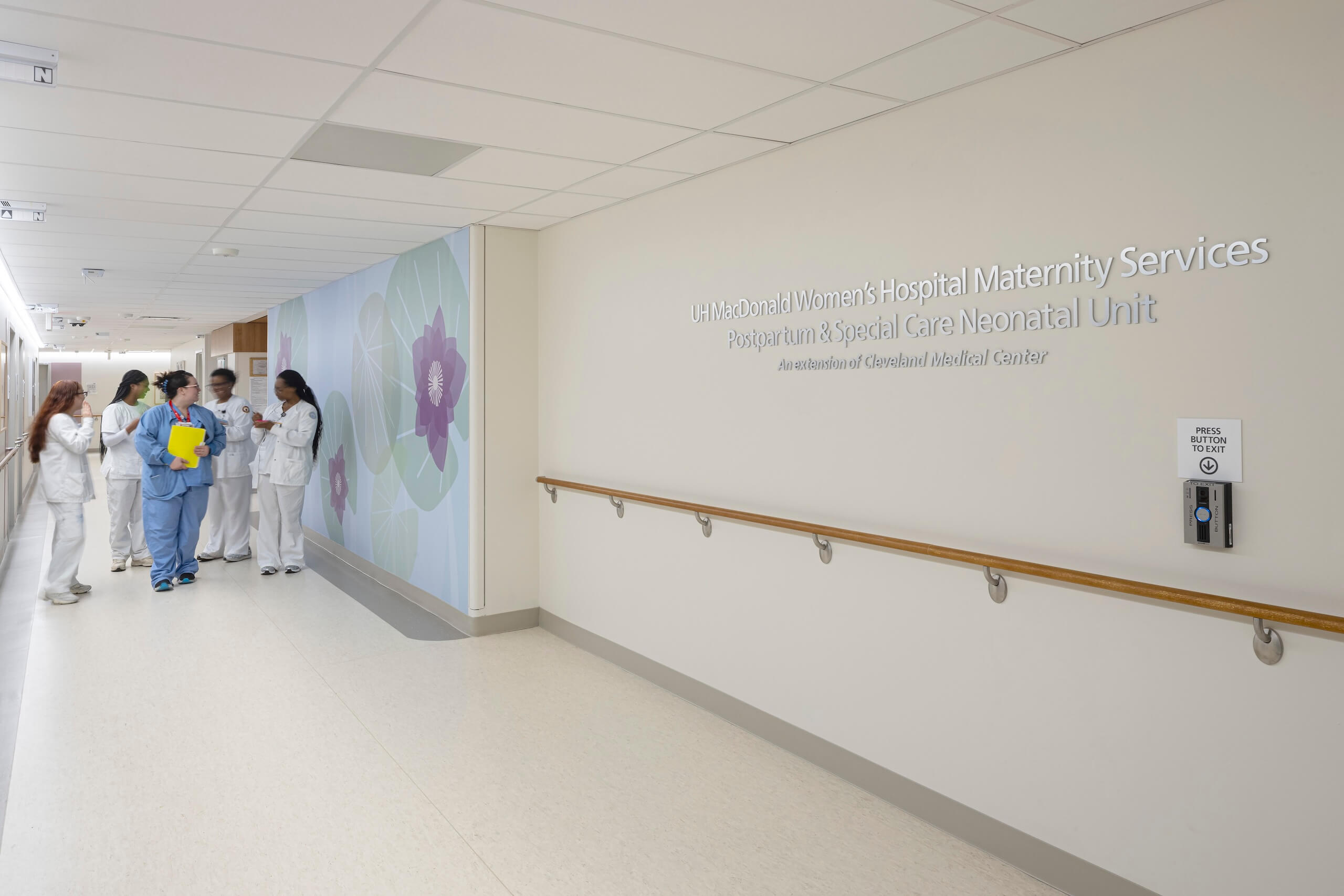 A group of healthcare workers in scrubs stand talking near a hospital corridor. A wall sign reads Maternity Services and Neonatal Unit next to a handrail and an accessible door button.