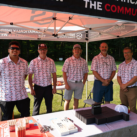 Five men in matching patterned shirts stand and smile under a canopy at an outdoor event table, with promotional items and a wooded area in the background.