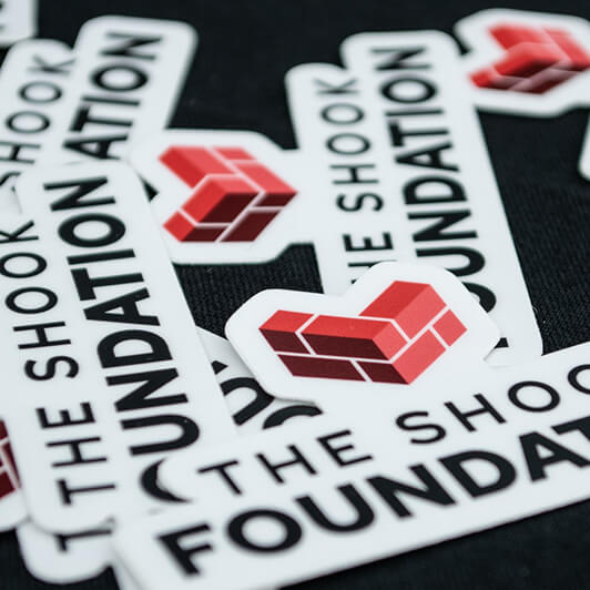 Several stickers featuring the words THE SHOOK FOUNDATION and a red-brick-style heart logo are scattered on a dark surface.