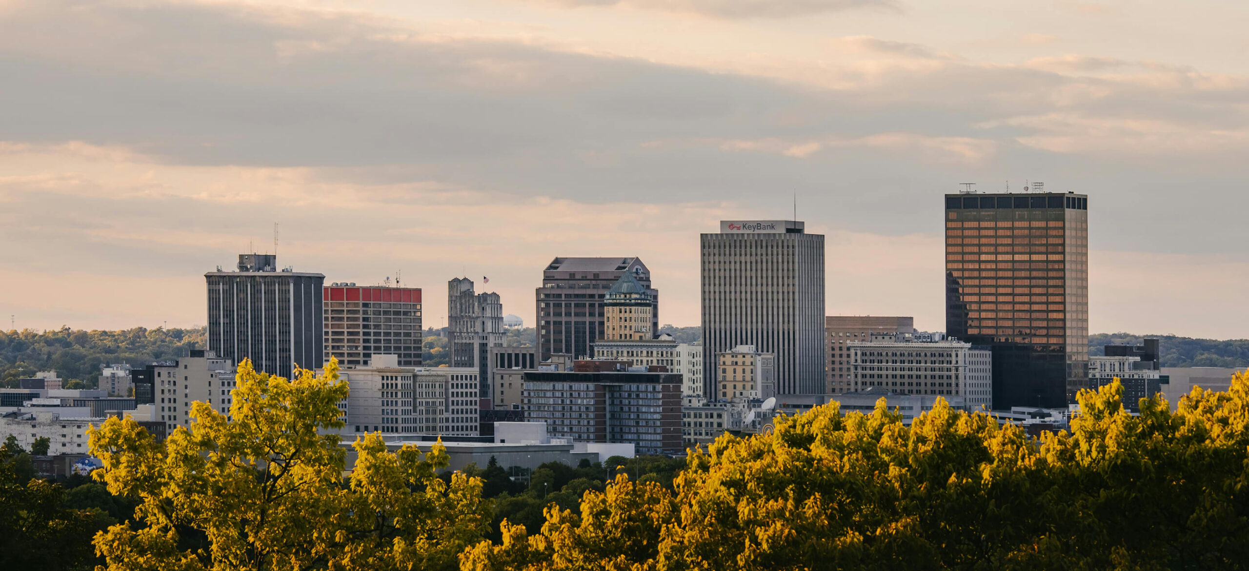 A city skyline with mid-rise and high-rise buildings, seen behind green and yellow leafy trees under a partly cloudy sky at sunset.