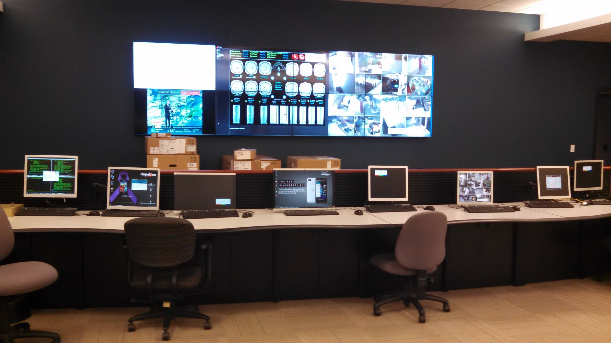 A control room with seven workstations, each with a monitor and keyboard. Multiple screens on the wall display surveillance cameras, data, and maps. Cardboard boxes are stacked beneath the large screens.