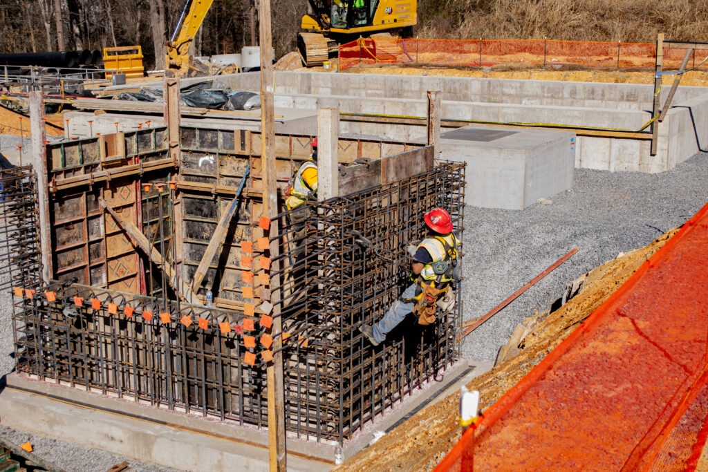 Two construction workers in safety gear work on a reinforced steel structure at a building site, surrounded by concrete forms, construction equipment, and safety barriers on a sunny day.