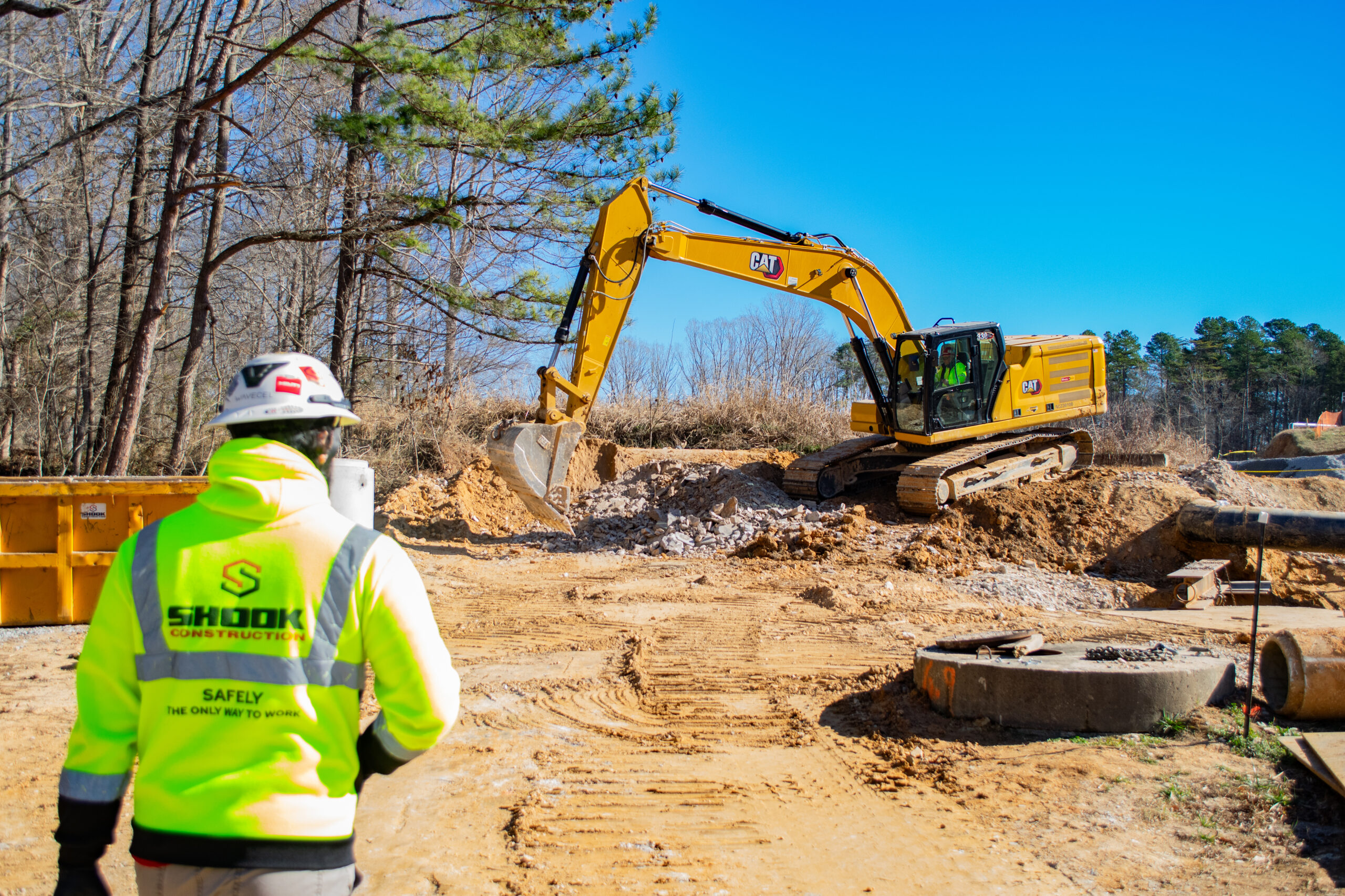 A construction worker in a neon jacket and helmet observes a yellow excavator digging at a sunny construction site with trees in the background.